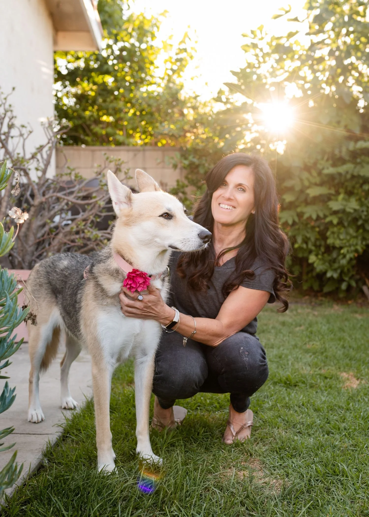 A woman with long dark hair kneels on the grass, smiling and holding a large husky dog with a pink flower collar outside in a garden during sunset.