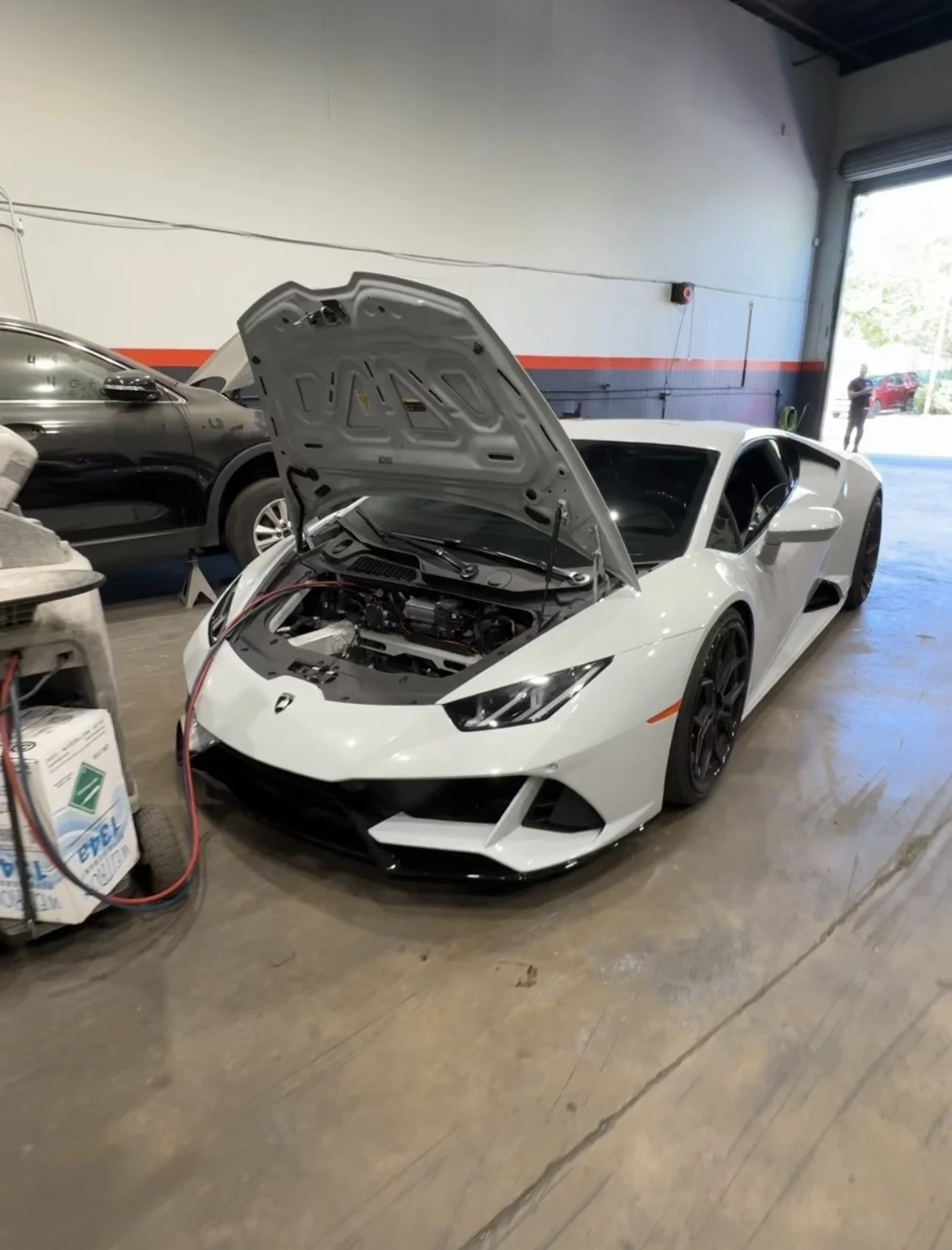 A silver Lamborghini sports car is parked inside a garage with an automatic car wash machine nearby. Outside, a few other cars and a tree are visible through the open garage door.