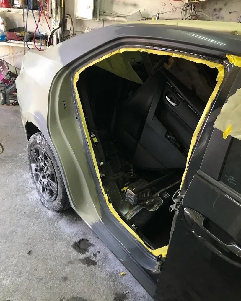 The interior of a car being repaired, with the driver's side door and window removed and masking tape around the opening, in a garage with tools and equipment in the background.