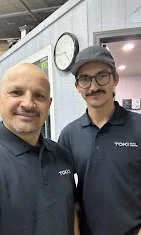 Two employees (Father & Son) wearing dark blue shirts taking a selfie indoors, with a clock on the wall behind them.