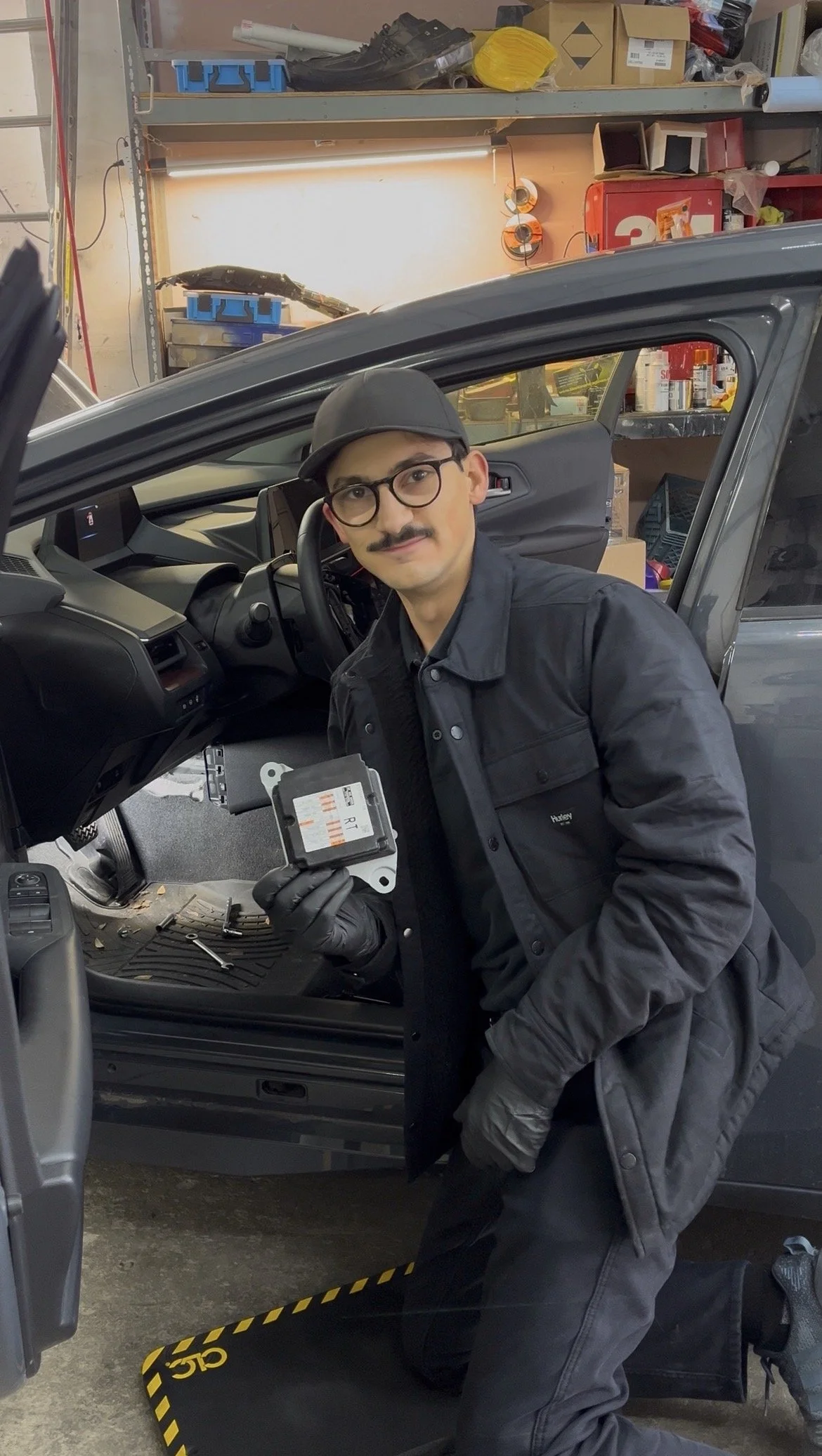 A technician, kneeling next to a car in a workshop, holding an airbag module with tools nearby.