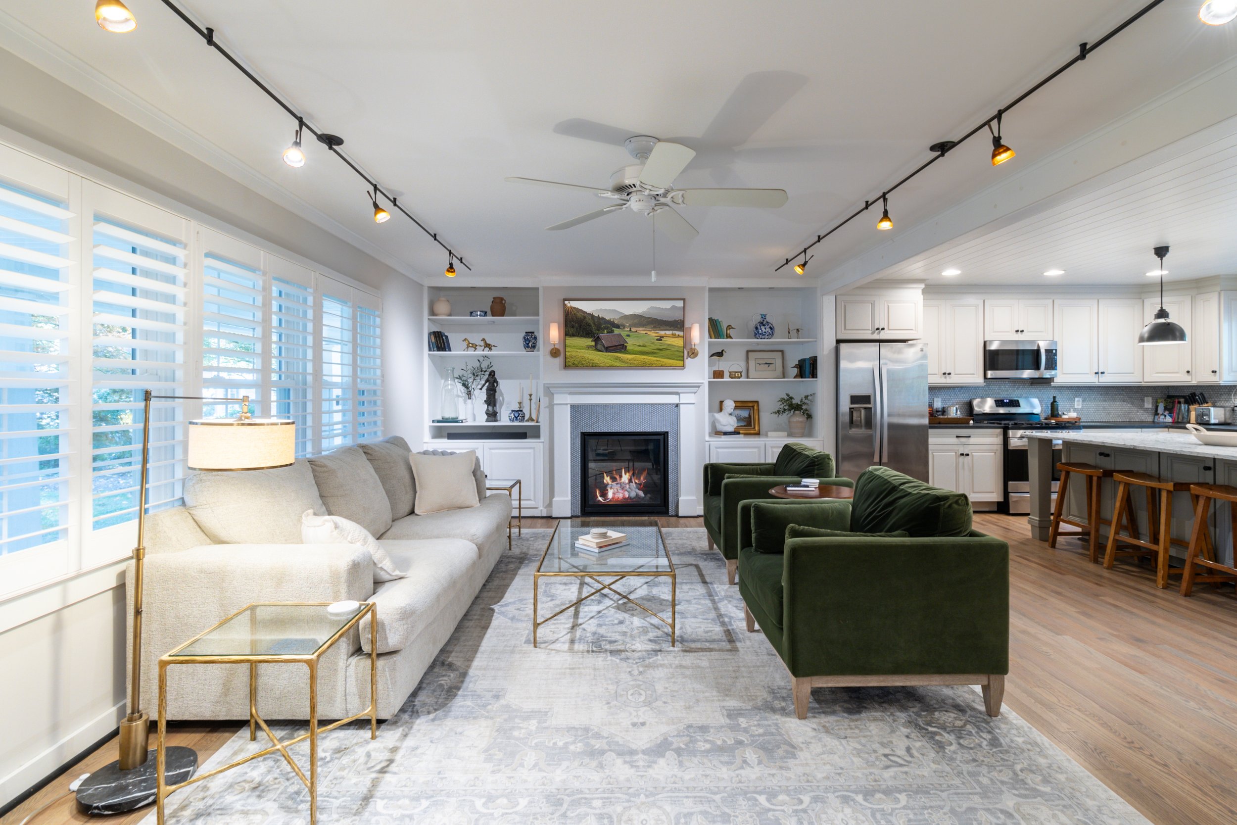 Living room with large windows, white sofa, three green armchairs, a fireplace with a painting above, built-in bookshelves, a TV, and a kitchen with white cabinets, stainless steel appliances, and a breakfast bar with wooden stools.