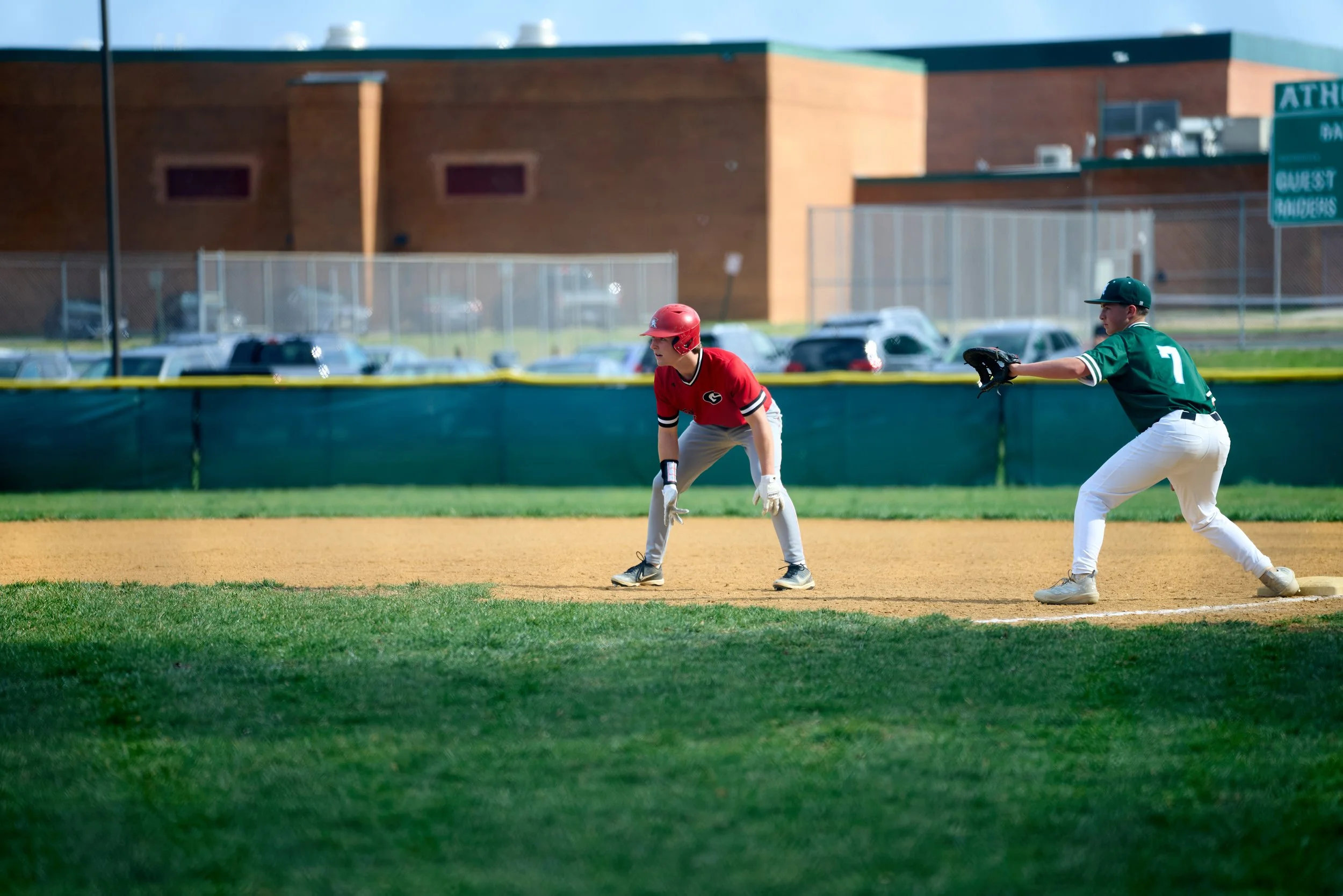 Two young baseball players on the field, one in a red uniform and the other in a green uniform, with a sports complex and parking lot in the background.