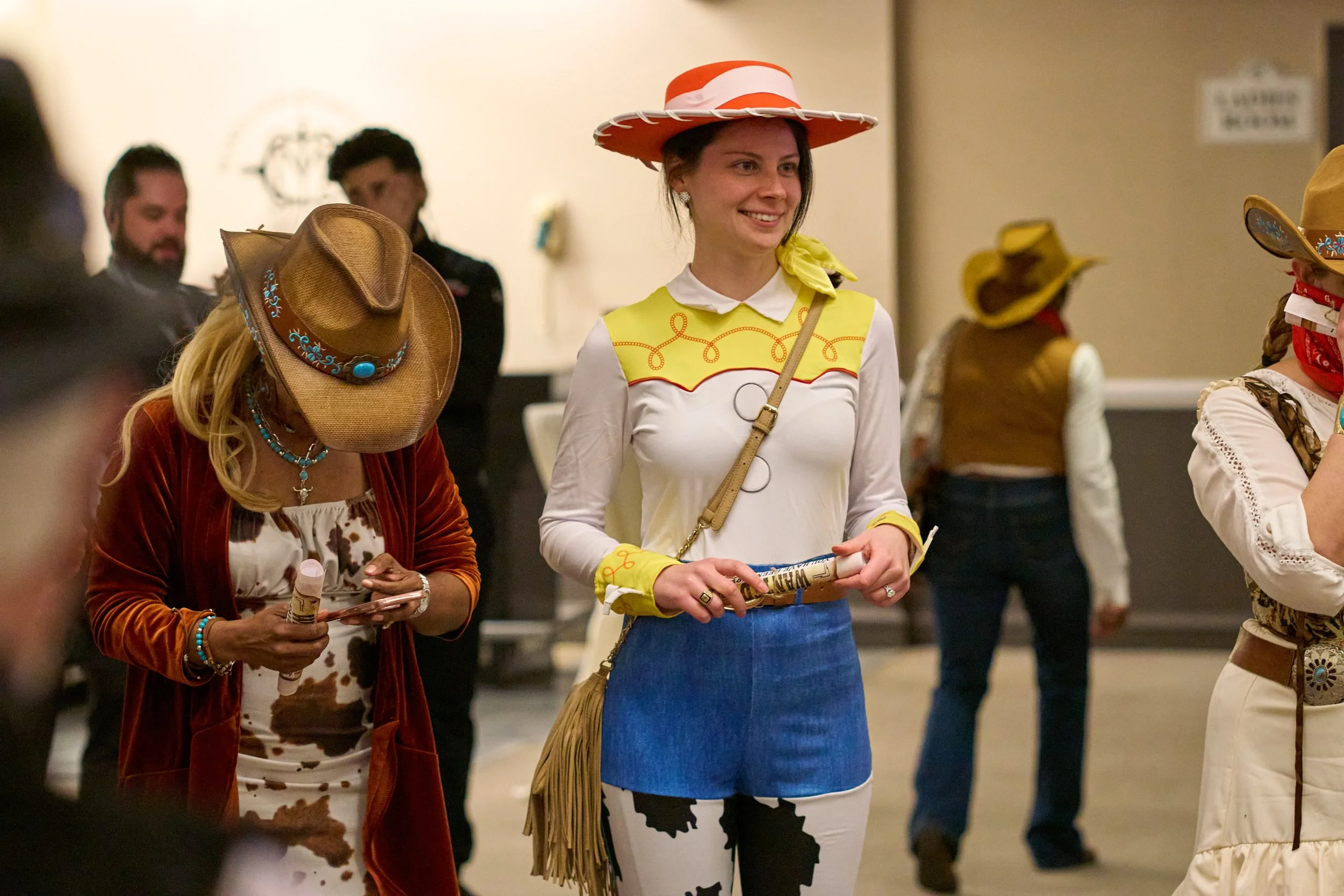 People at a themed event wearing cowboy and cowgirl costumes, including hats and western-style clothing, with some holding scrolls or other objects.