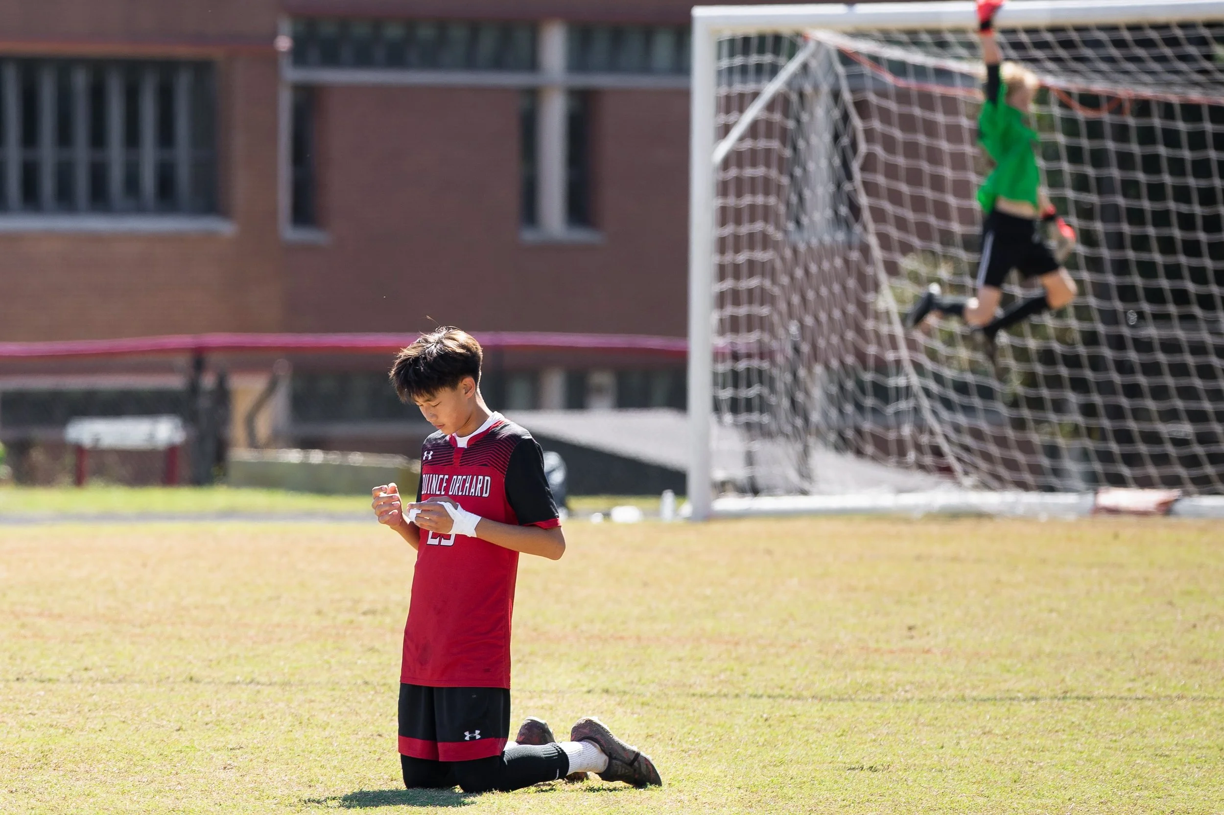 A boy kneeling on a soccer field, holding his hands together, with a soccer goal and goalkeeper in the background.