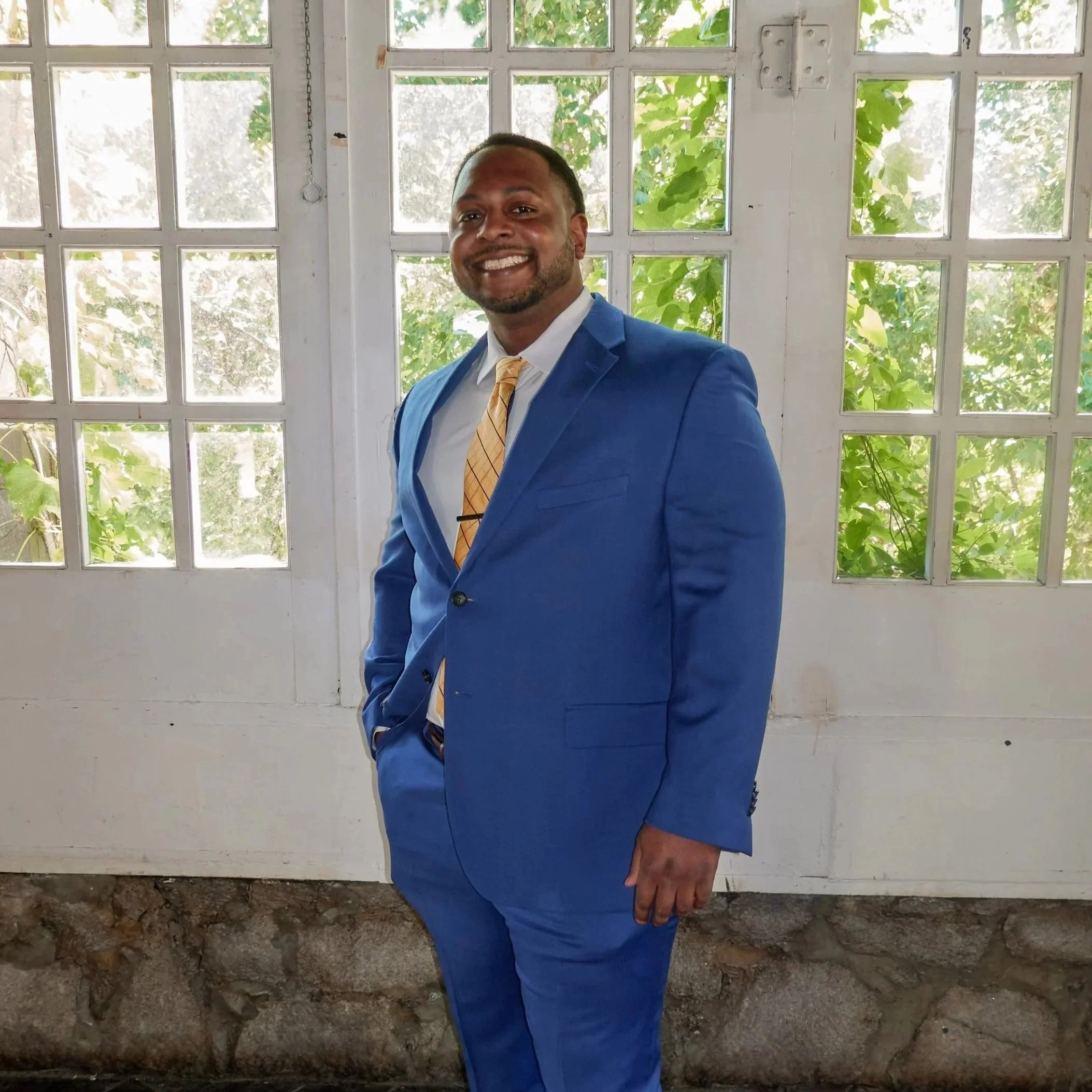 A man in a blue suit, white shirt, and yellow plaid tie posing indoors in front of a window with a stone and wood wall beneath.