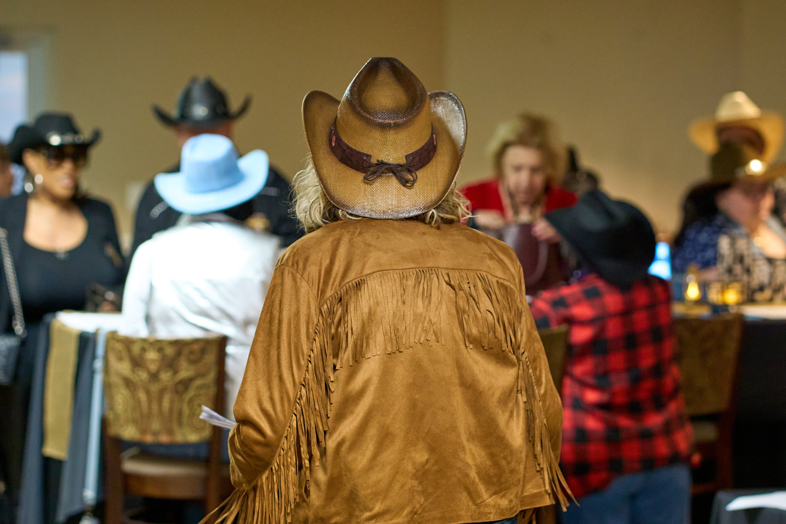 A woman in a brown fringed suede jacket and a brown cowboy hat is seen from behind, with a group of people wearing cowboy hats and western-style clothing in the background.
