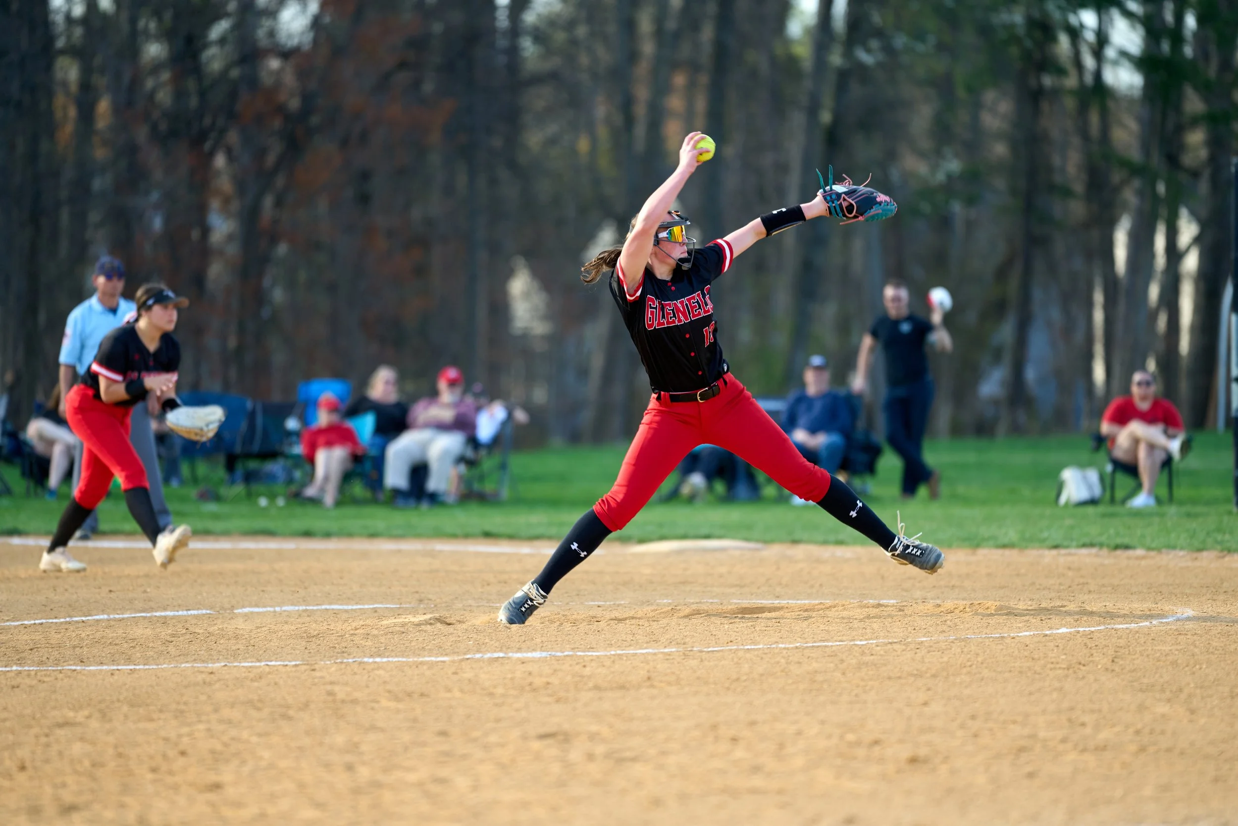 A female softball pitcher in a red and black uniform throwing a pitch on a softball field. She is wearing glasses and her hair is tied back. In the background, there are spectators seated in chairs and trees.