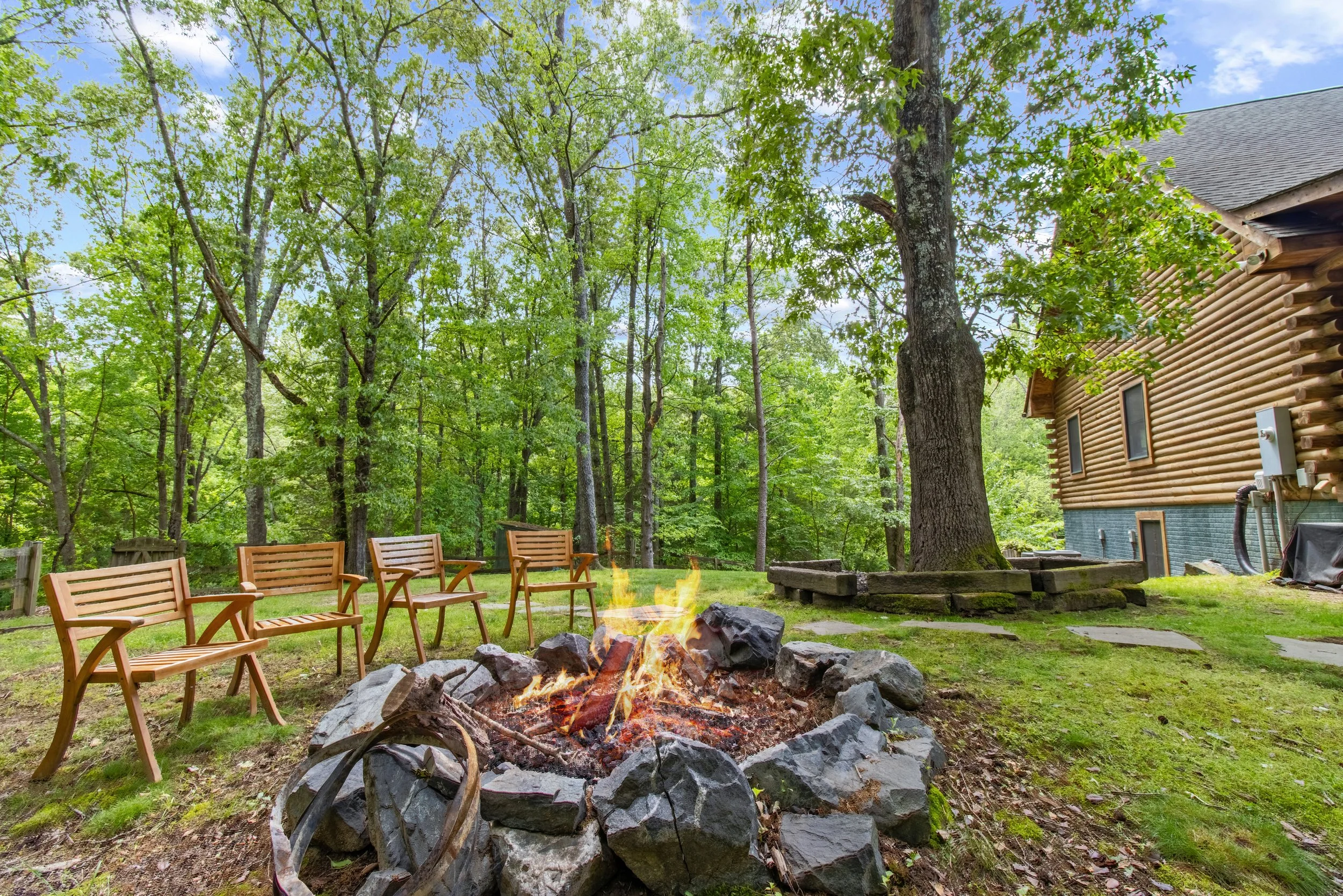 A cozy outdoor fire pit with flames, surrounded by five wooden chairs, situated on a grassy area in a wooded backyard with tall trees and green leaves. To the right is a log cabin-style house with siding and small windows.