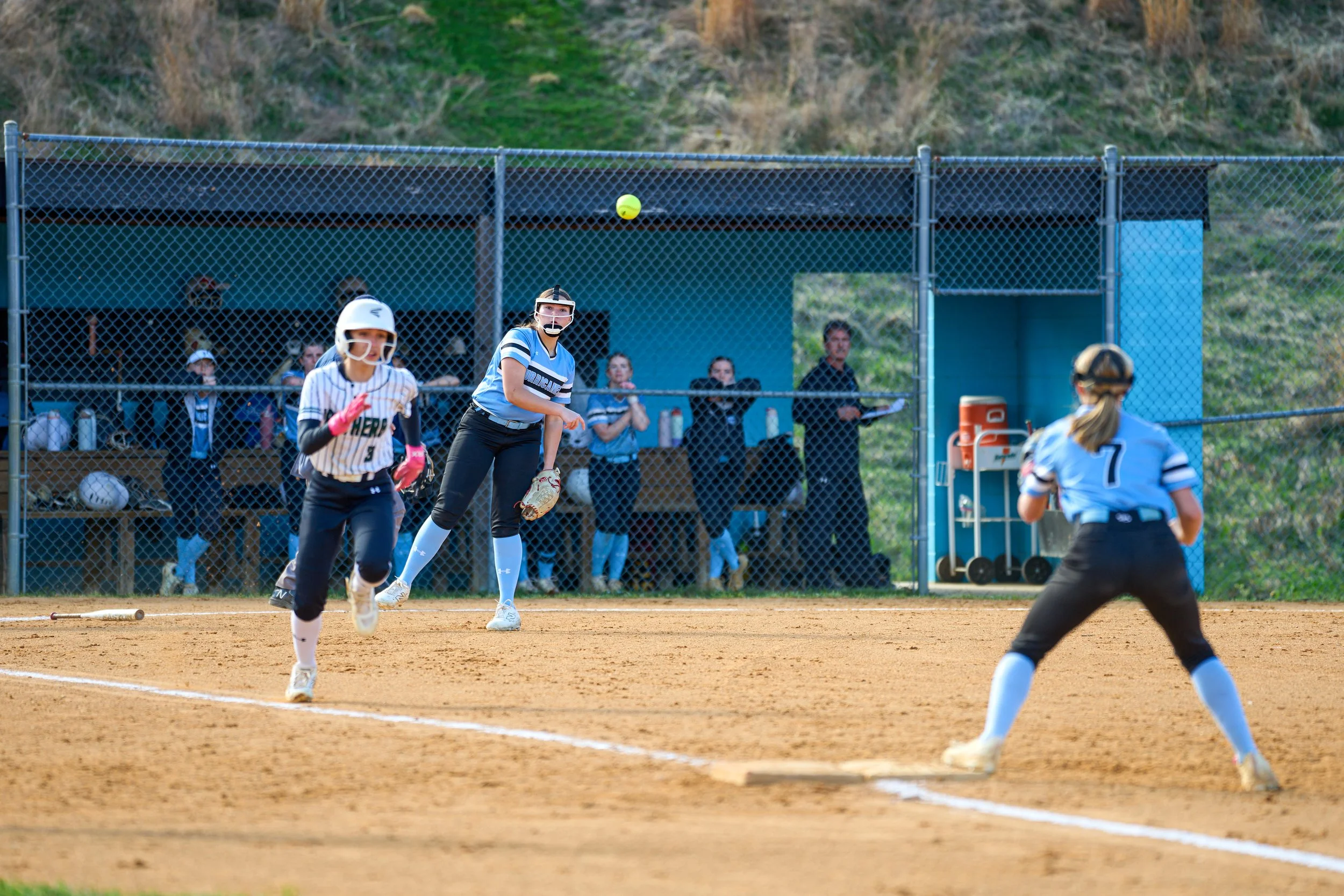 Softball game with players running on the field and a player in the act of catching a ball. The infield is dirt, with a fence and spectators in the background.