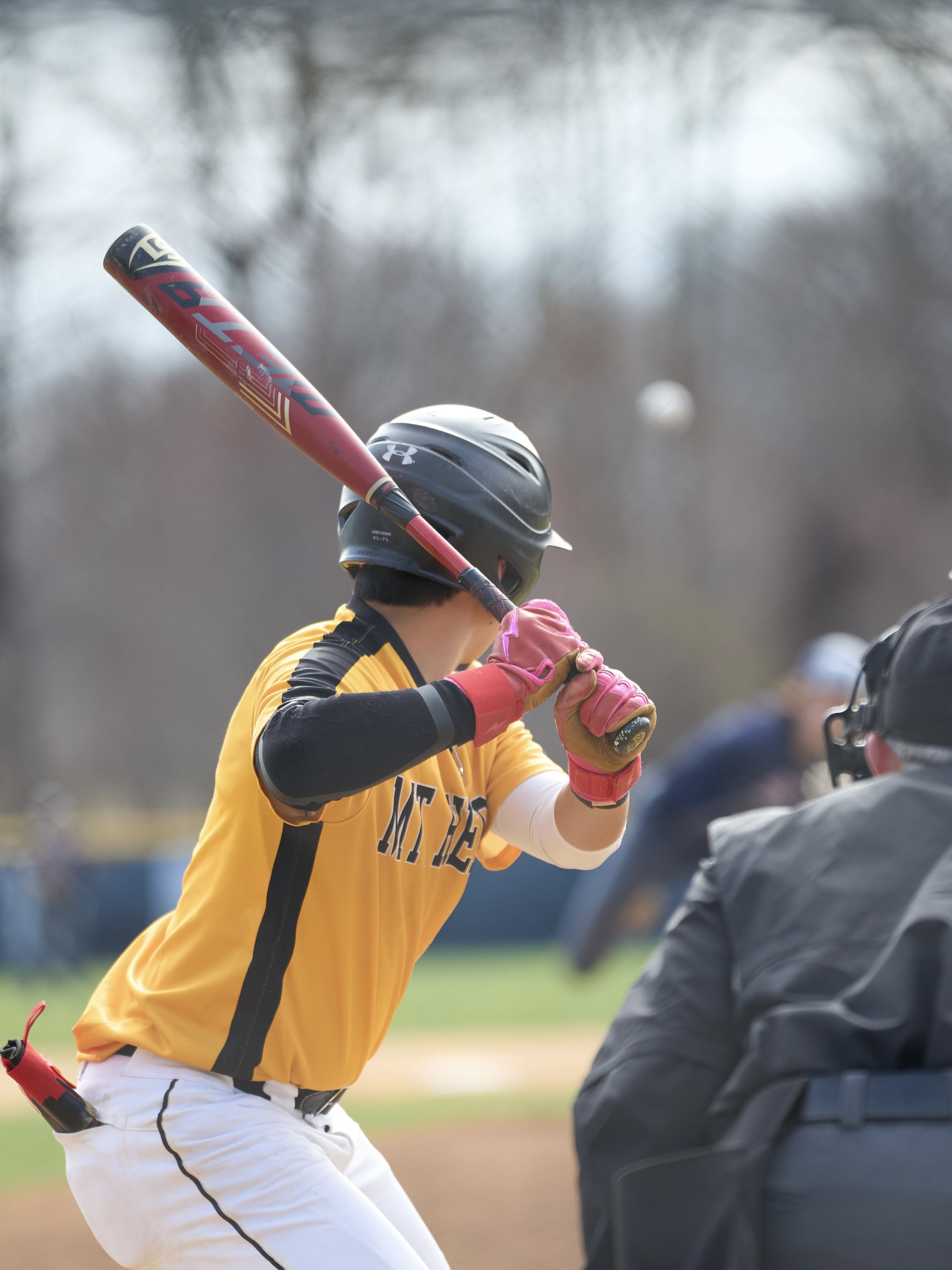 Baseball player in yellow uniform preparing to hit with bat, wearing black helmet and pink gloves, on field with umpire nearby.