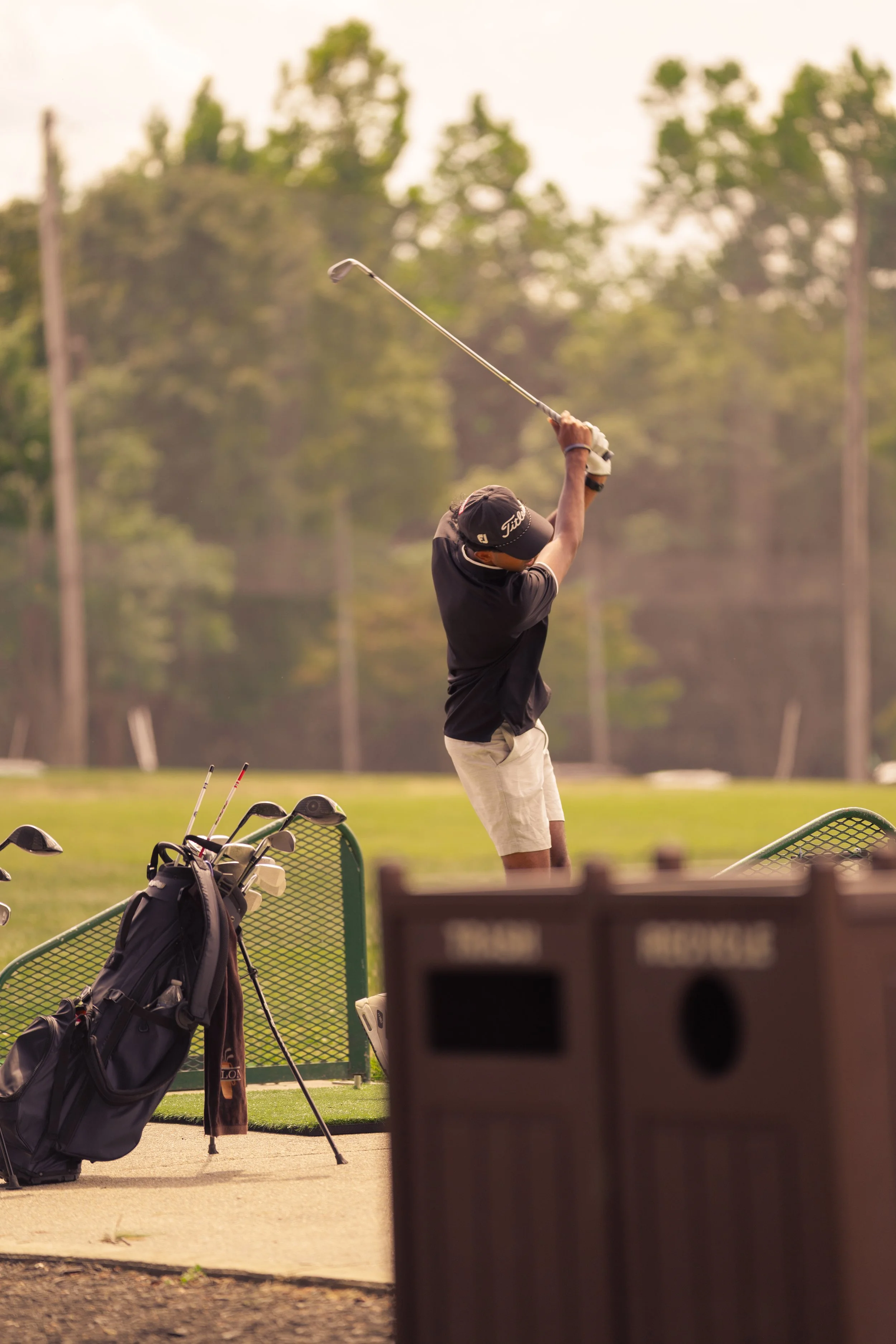 A person playing golf taking a swing on a golf course. The golfer is dressed in a black shirt, beige shorts, and a black cap, with a golf bag and clubs nearby.