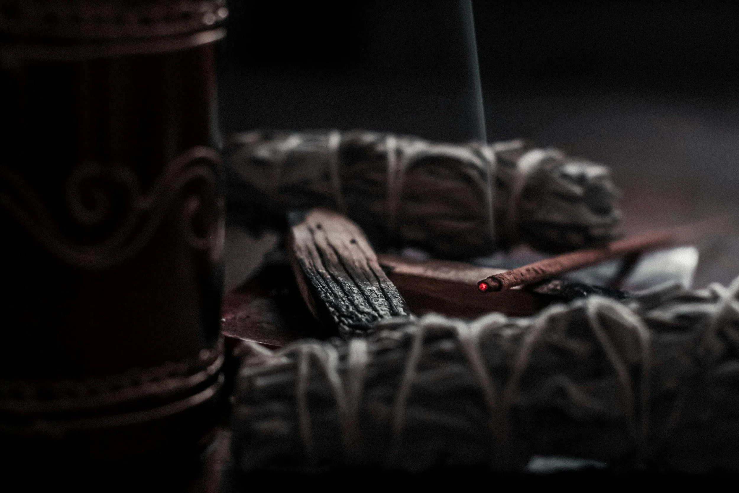 A partially lit still life scene with a wooden cigar box, charred wood, a lit cigarette, and a ring of white string on a dark surface.