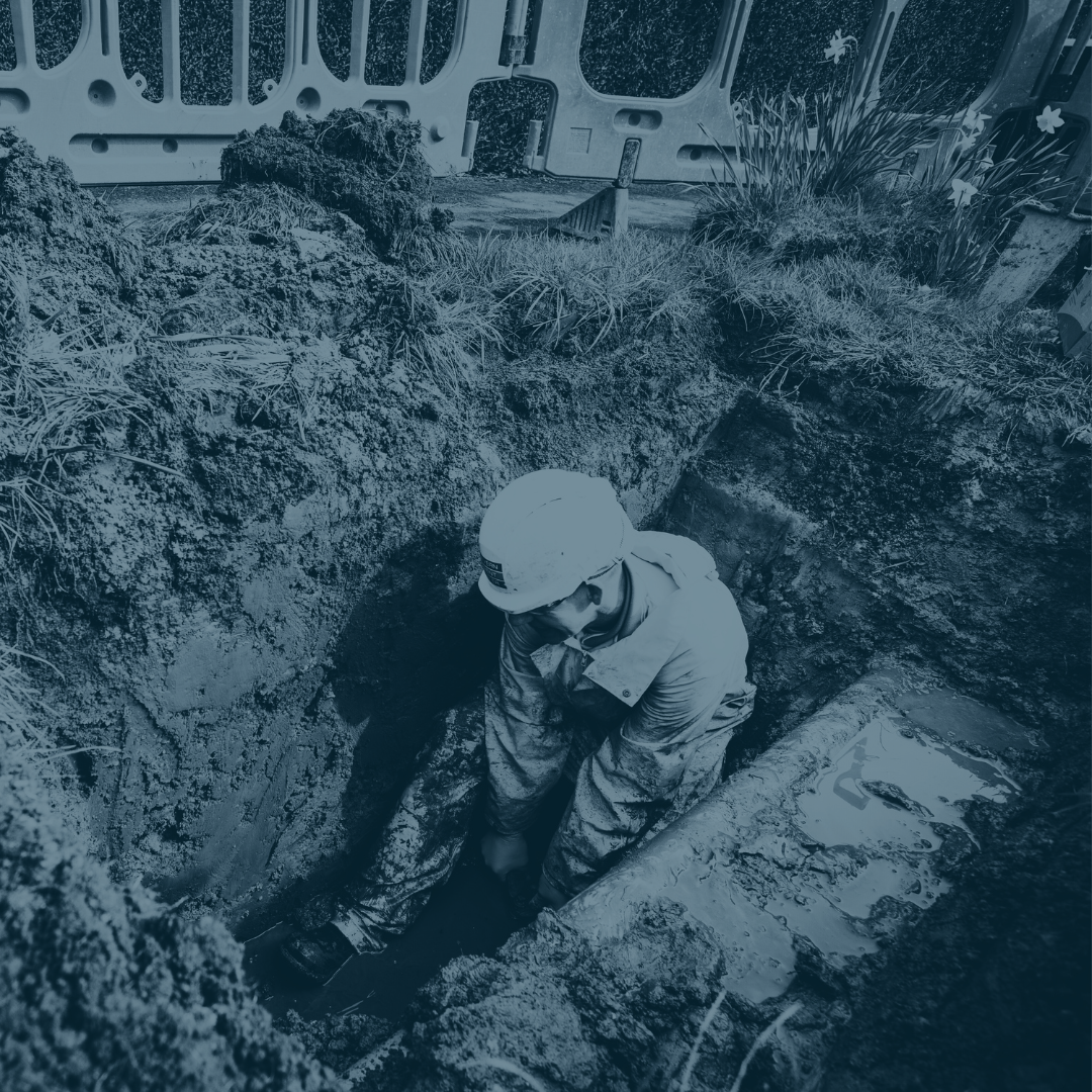 A construction worker wearing a hard hat and safety gear crouches inside a deep trench at a construction site.