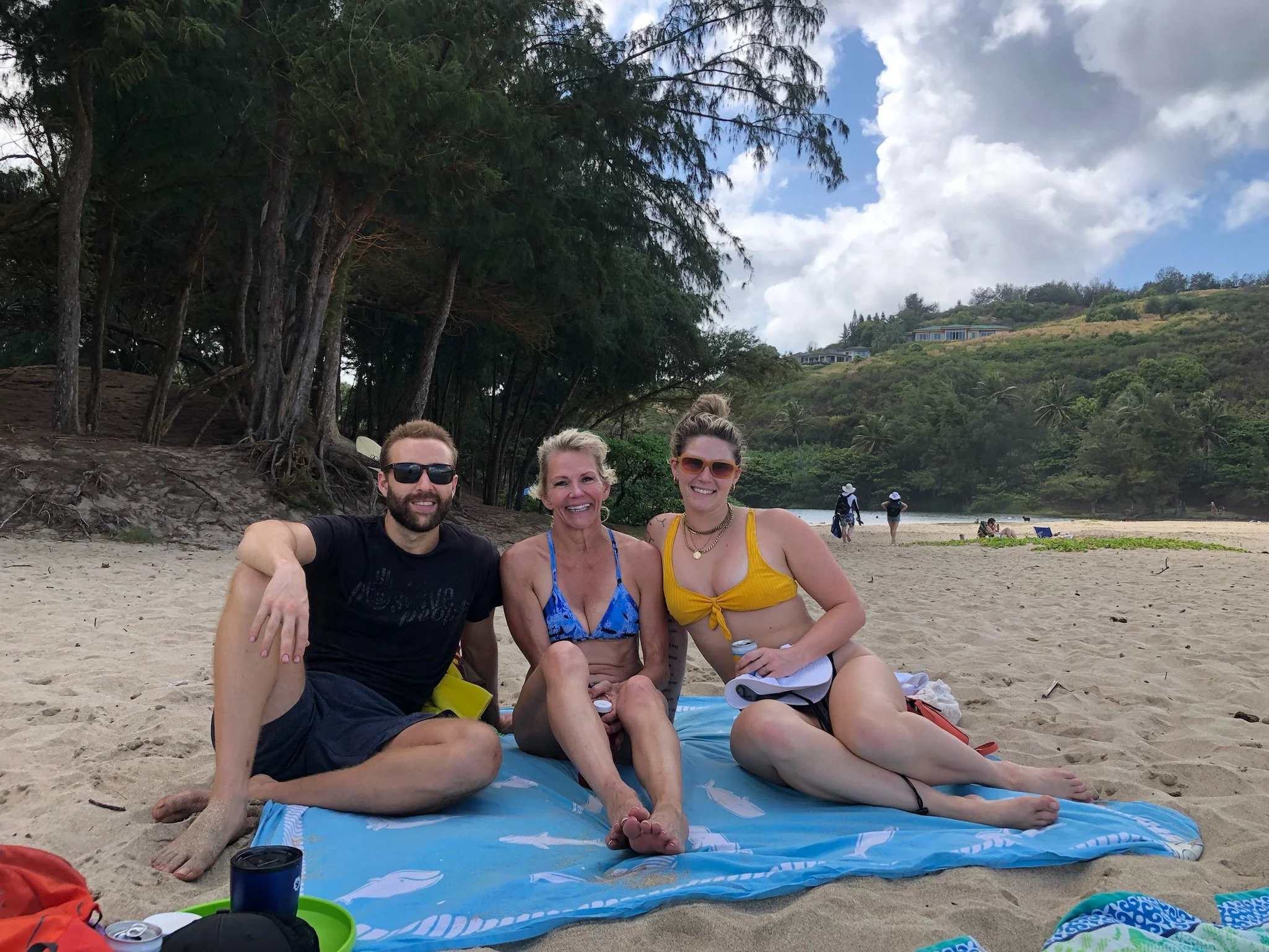 Four smiling people sitting on a towel on a sandy beach, with palm trees, hills with houses, and partly cloudy sky in the background. Two women and one man are facing the camera, while the third person in the back is walking away.