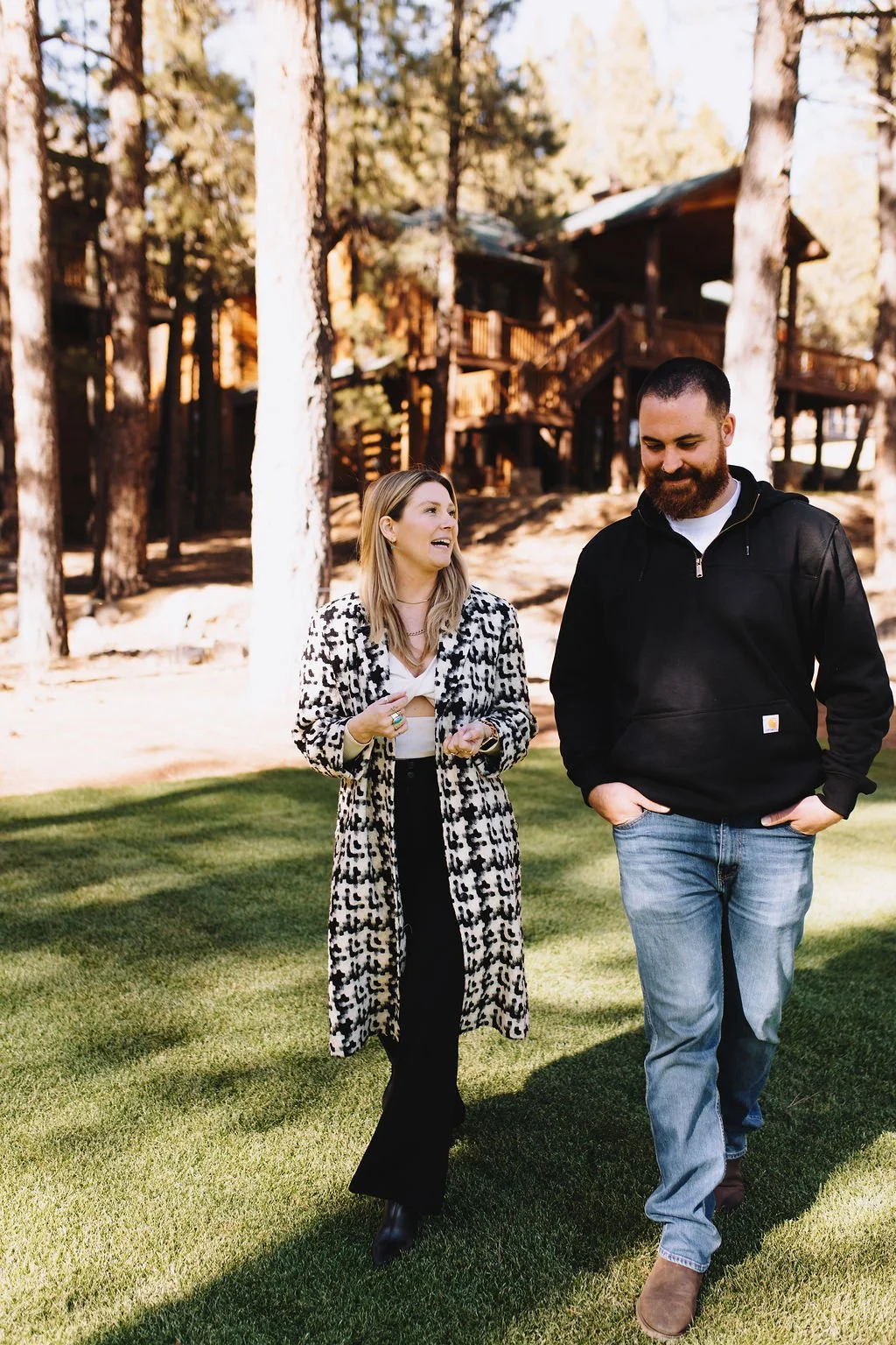 A woman and a man walking on grass in a wooded area with tall trees and wooden cabins in the background. The woman is wearing a leopard print coat and black pants, and the man is wearing a black hoodie and jeans.