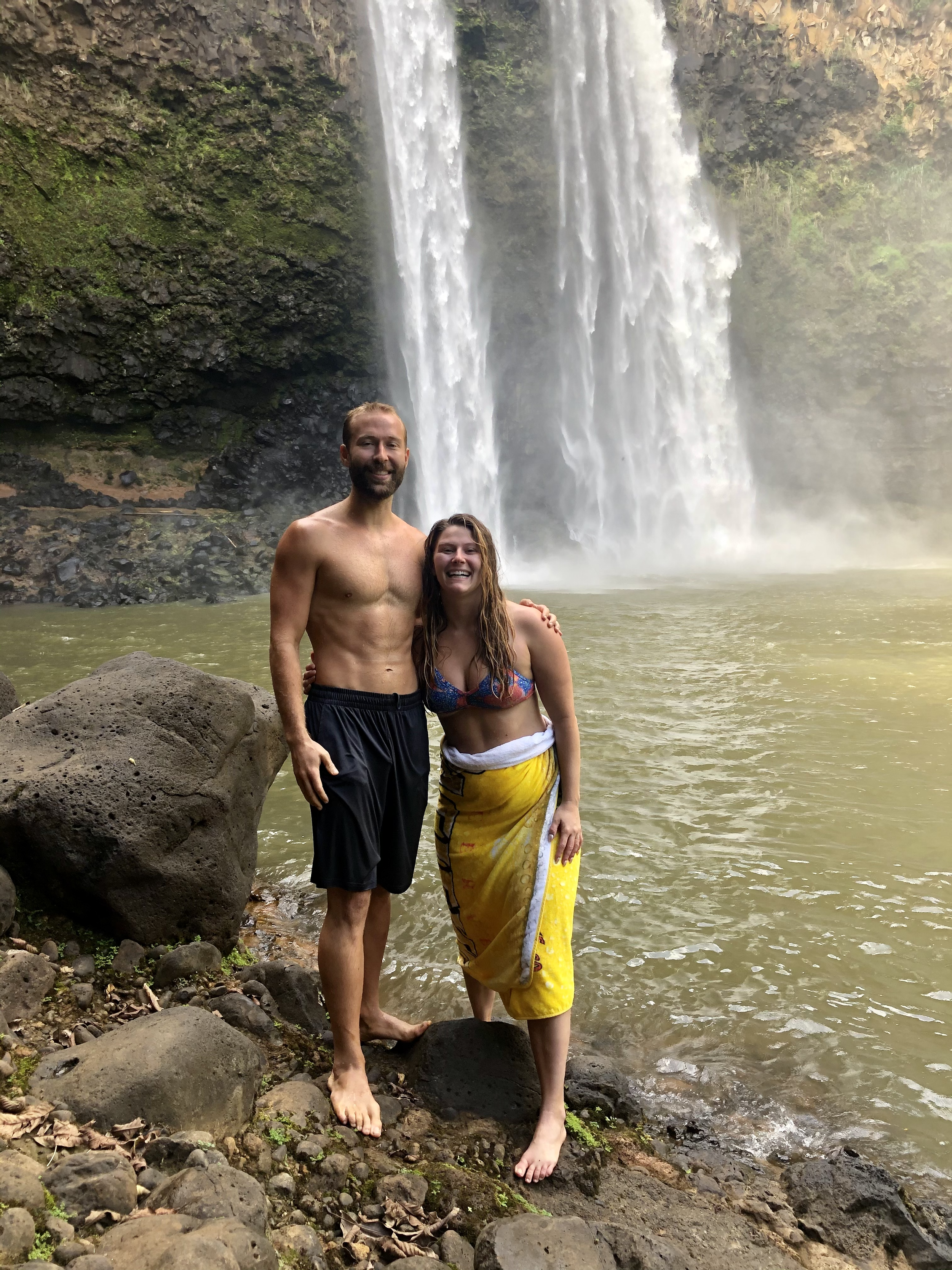 A man and a woman standing on rocks by a waterfall, smiling. The man is shirtless, and the woman is wearing a colorful bikini top and a yellow sarong. Water and greenery are visible around them.