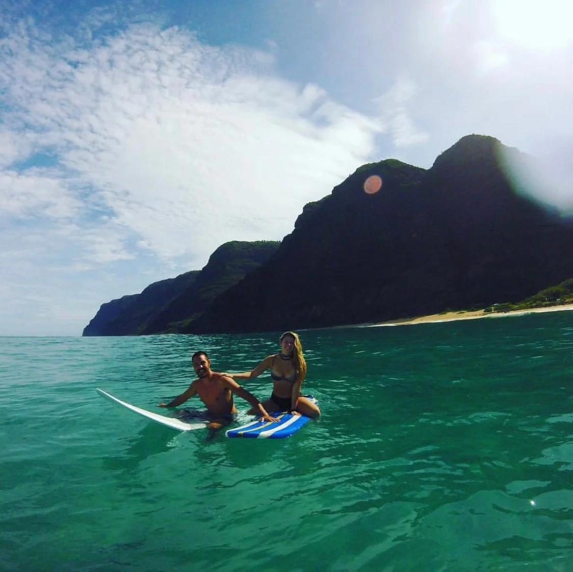 Two people surfing on a stand-up paddleboard in clear ocean water near a shoreline with large cliffs and a cloudy sky.