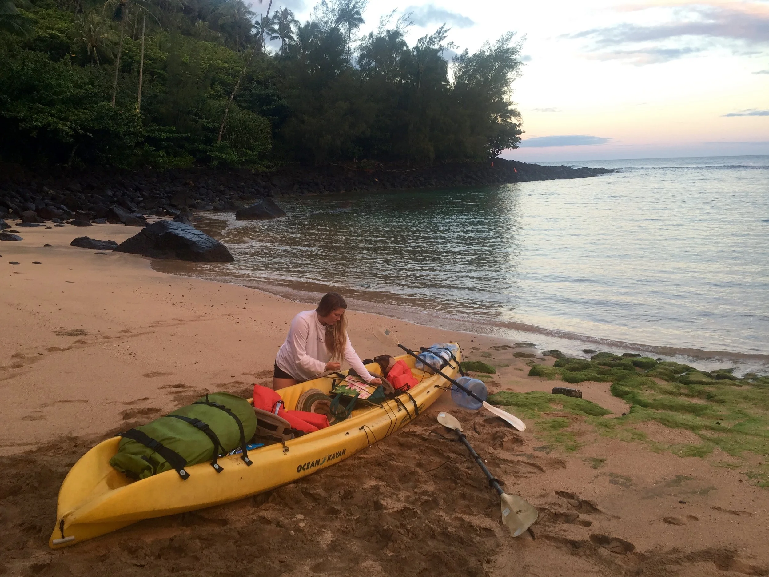 A woman preparing a yellow kayak on a sandy beach with trees and rocks, calm ocean water, and a partly cloudy sky in the background.