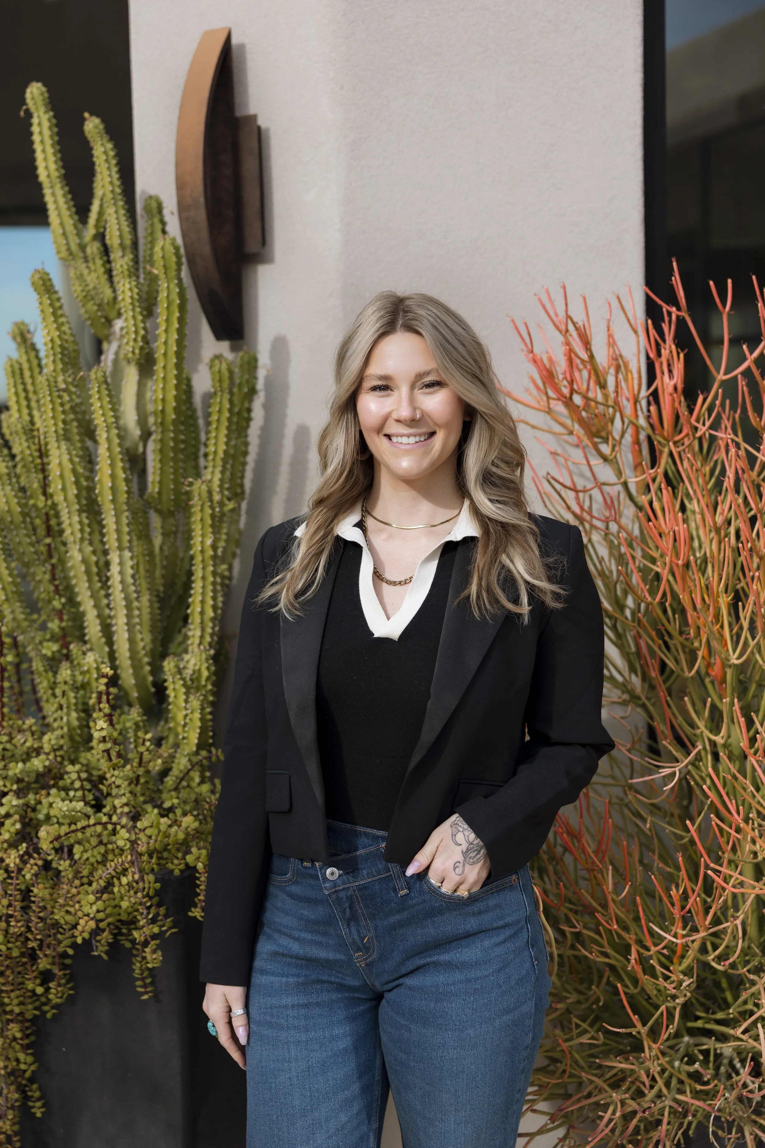 A woman with long wavy blonde hair, wearing a black blazer over a black sweater and blue jeans, standing outdoors next to cacti and succulents, smiling at the camera.