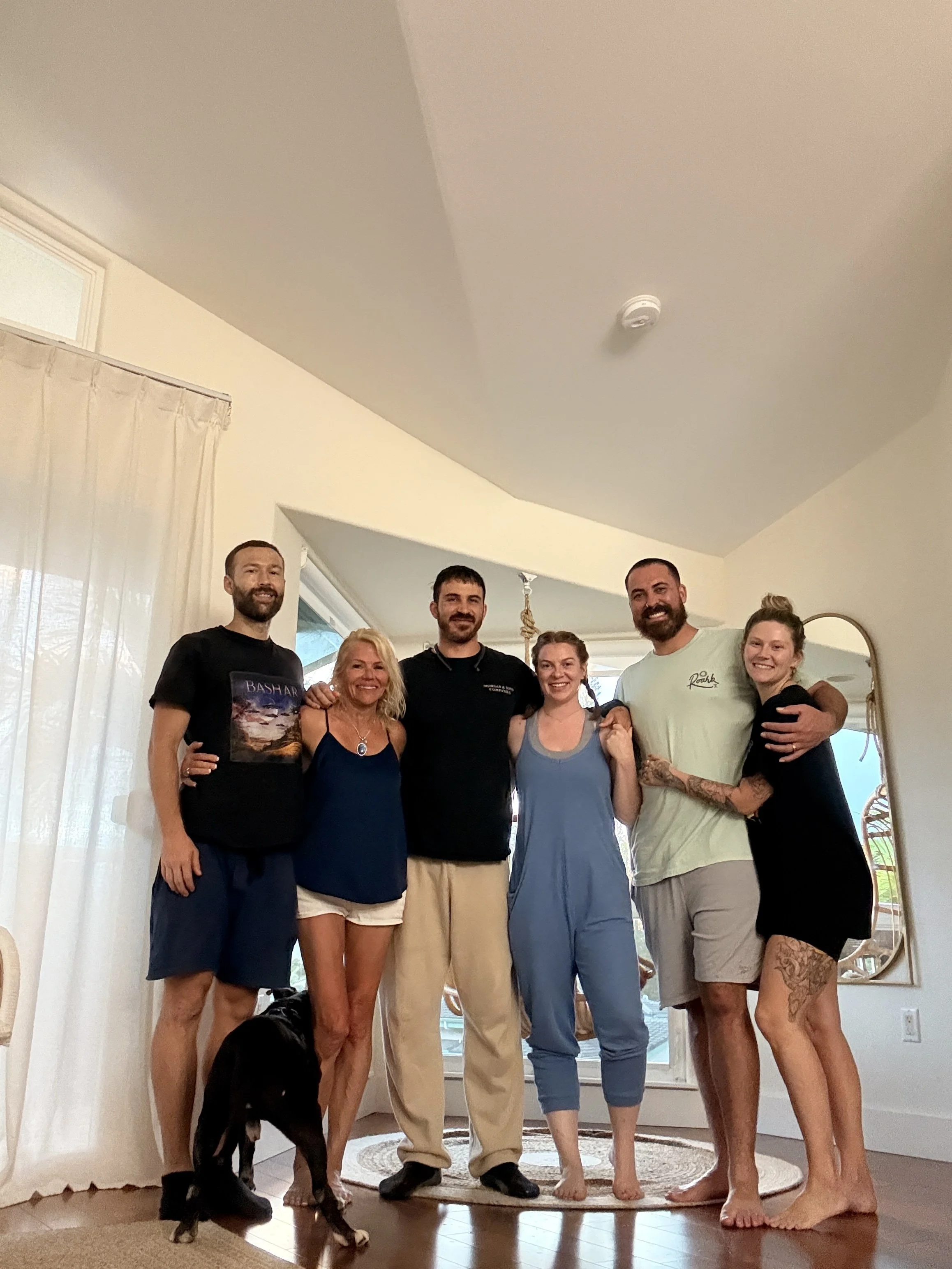 Group of seven people standing inside a home, smiling at the camera. They are standing on a round rug near a door with a large window, with curtains. There is a baby stroller on the floor and a black dog sitting in front of the group. The background shows a window with a view outside and part of a mirror on the right.