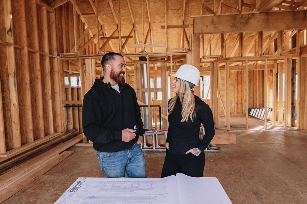 A man and woman in construction gear talking inside an unfinished building with wooden framing.
