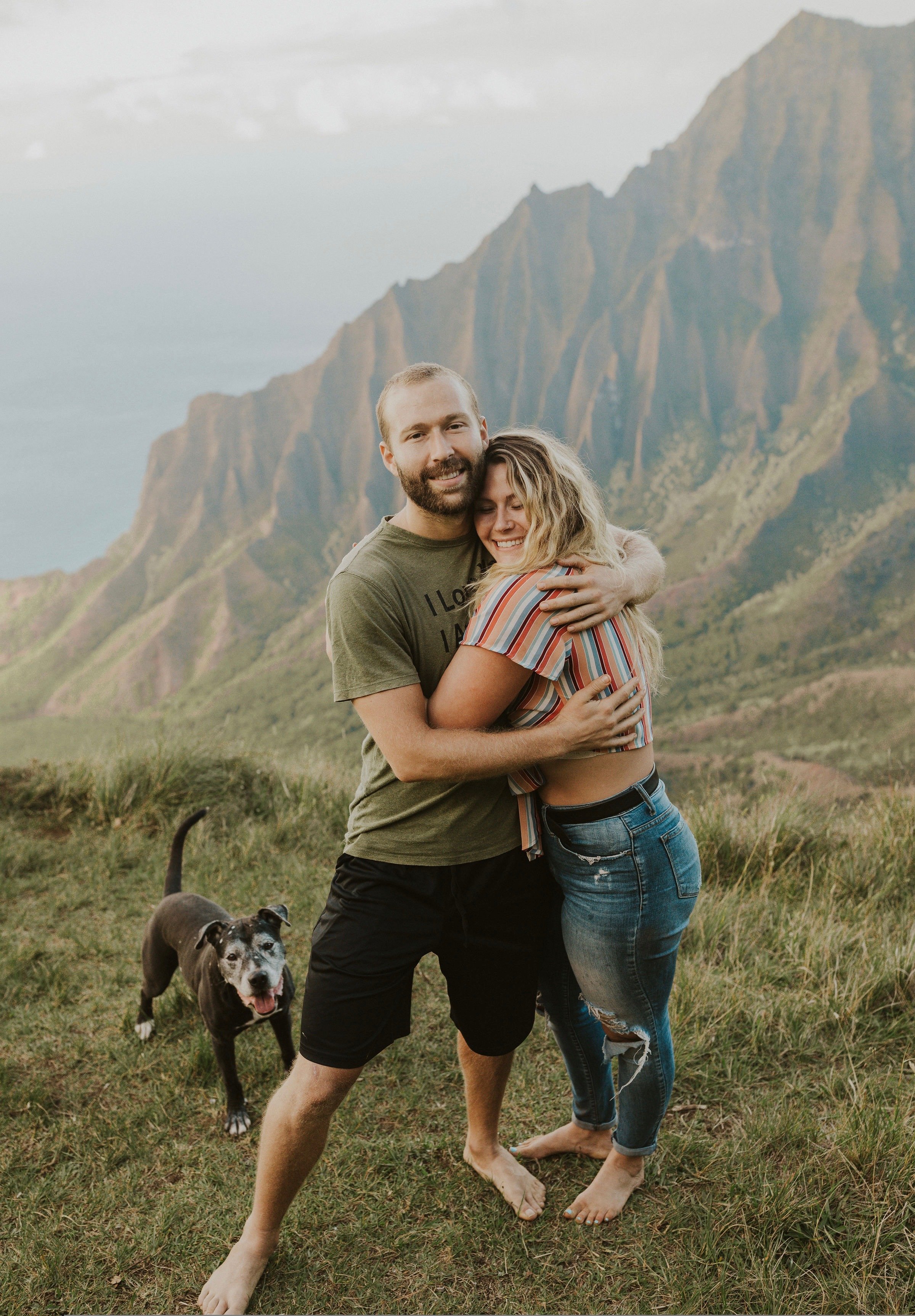 A happy couple hugging outdoors on a grassy field with a mountain range in the background. The man has a beard and is wearing a green t-shirt and black shorts. The woman has curly hair and is wearing a striped shirt and ripped jeans. A dog with a black and white coat is standing nearby, looking at the camera.