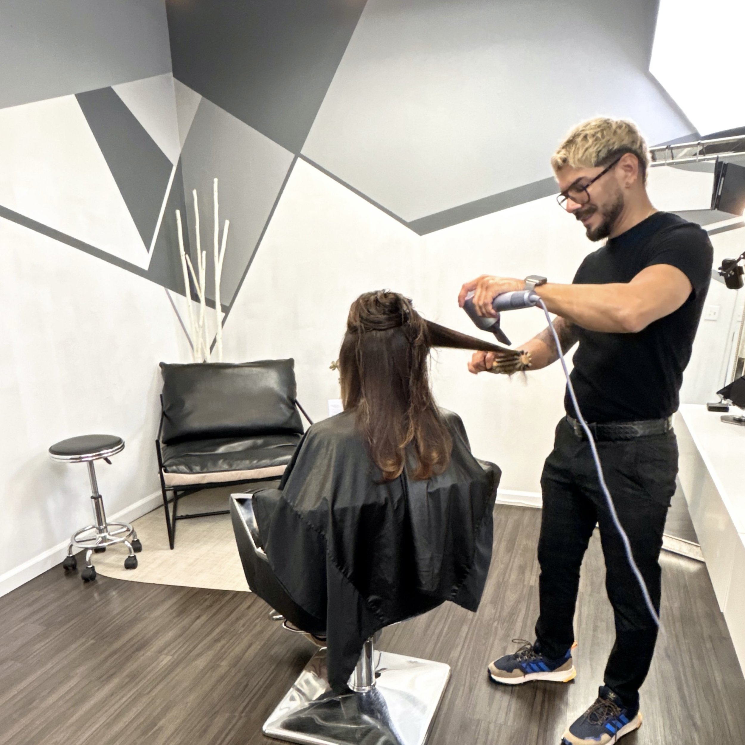 Hair stylist blow drying a woman's hair in a modern salon with geometric wall design.