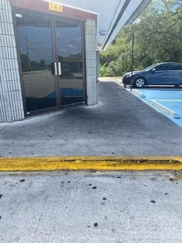 Empty sidewalk and parking lot with a black car parked in the distance. Building entrance with glass doors and a yellow parking space number sign above the door, yellow curb in the foreground. Trees and a partly cloudy sky in the background.