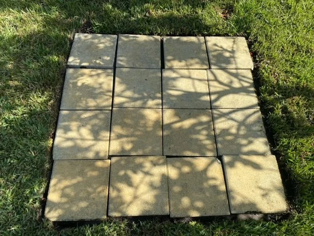 Square concrete paving stones arranged in a grid pattern on a grassy surface, with shadows of leaves cast across them.
