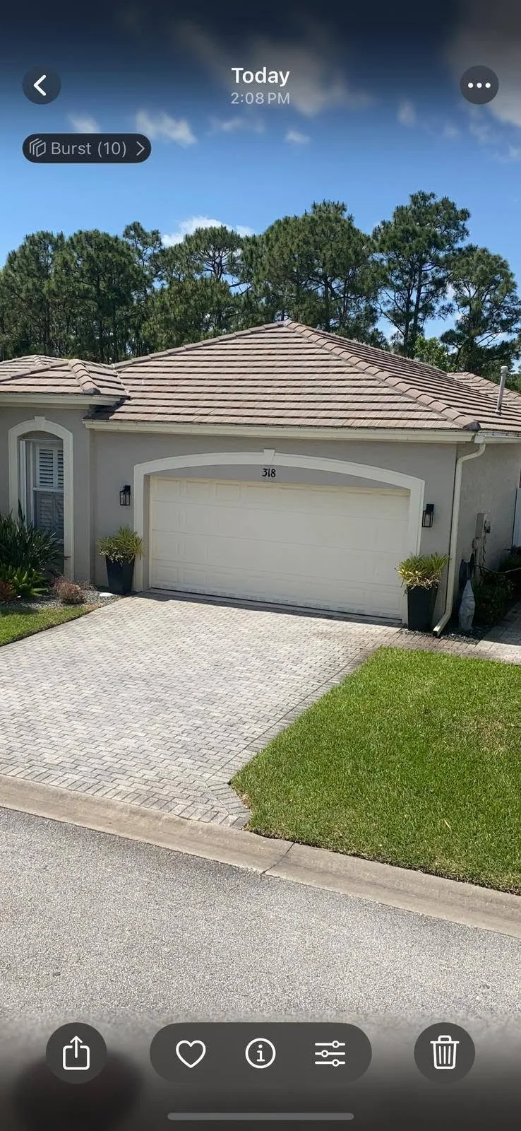 Front view of a single-story house with a white garage door, beige exterior walls, and a tiled roofing, with green plants on each side of the door and a paved driveway