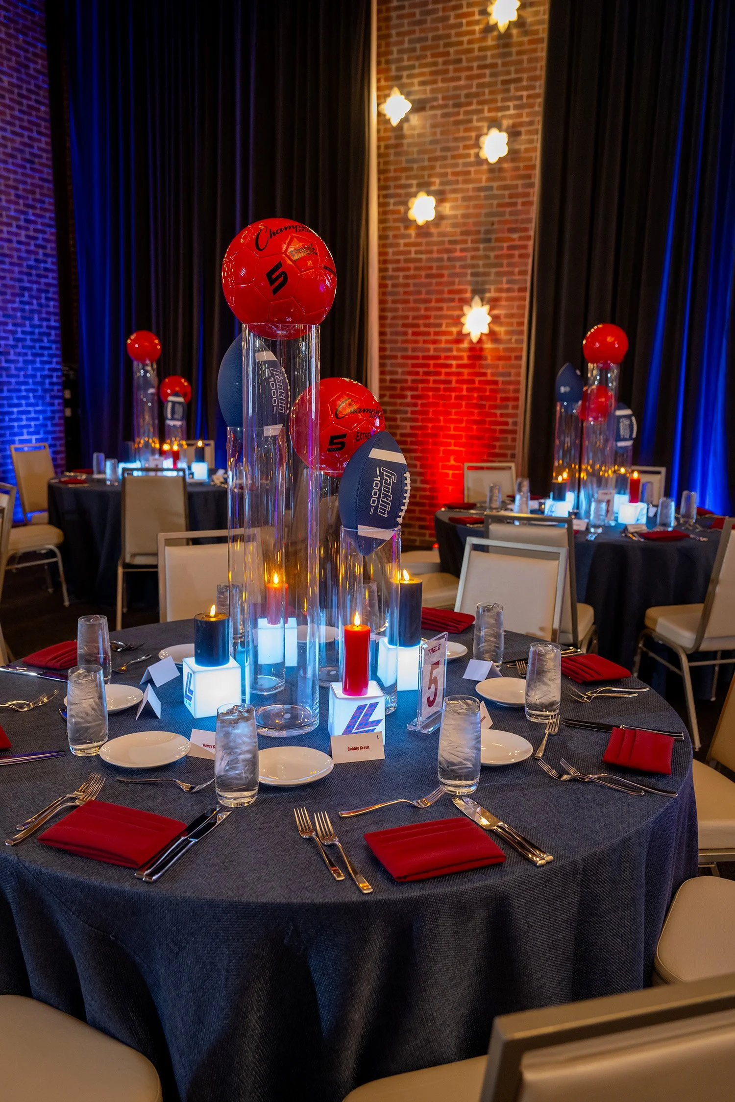 Round banquet table decorated with centerpieces featuring red and blue balloons and candles, set for a formal event with black tablecloths, white plates, silverware, and red napkins.