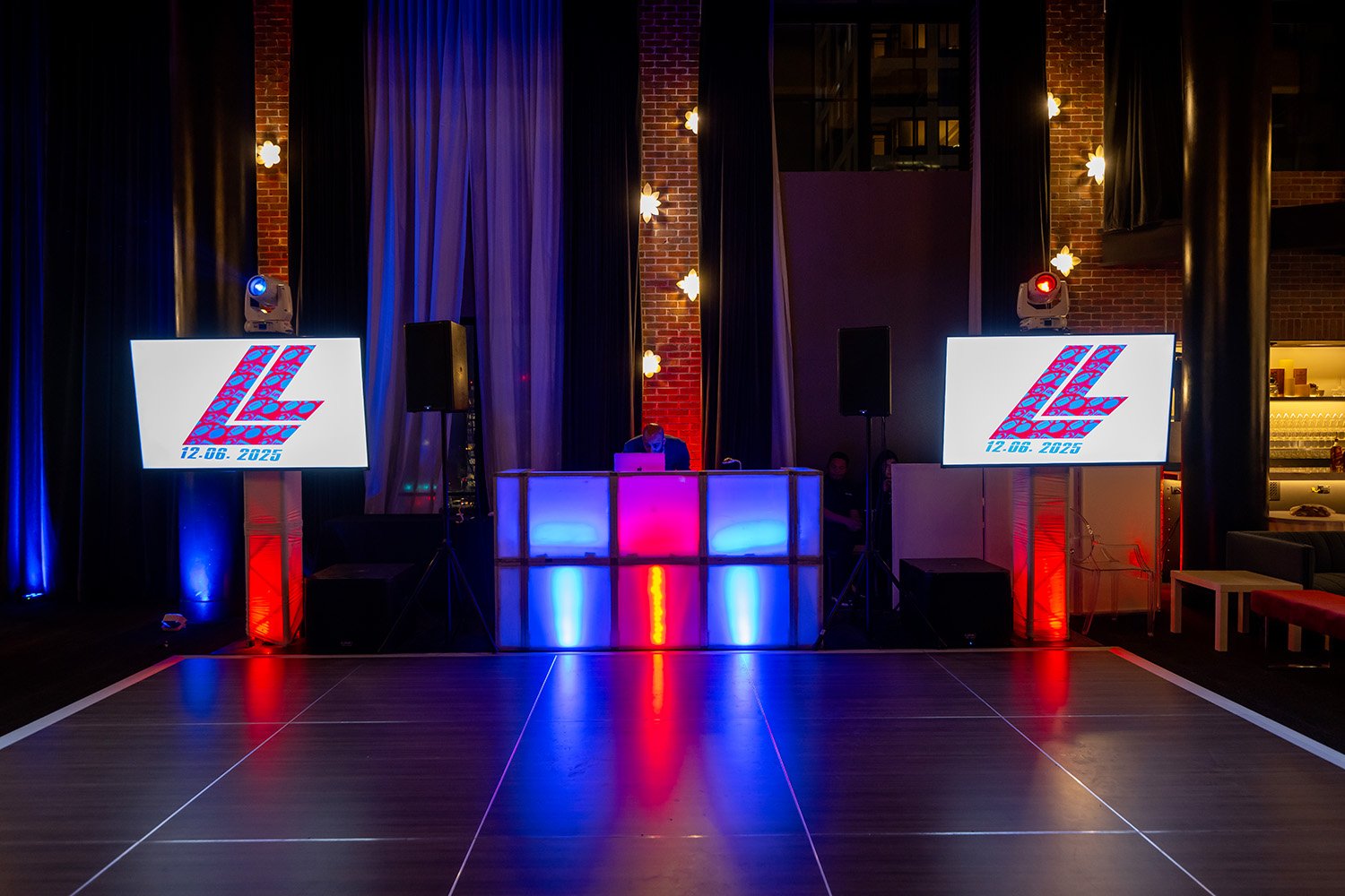 Bar or DJ setup with two large screens showing a stylized 'LZ' logo and date '12.06.2025', illuminated with red, blue, and purple lighting, located in a room with brick walls, curtains, and a dance floor.