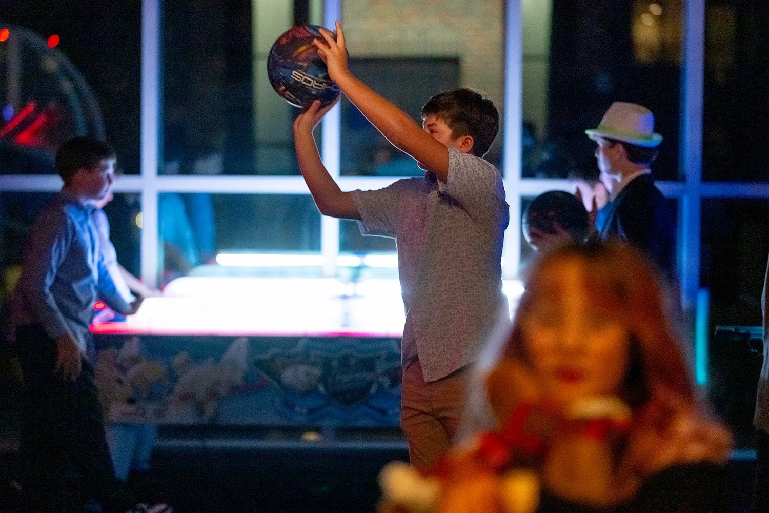A boy holding a hockey puck in a dimly lit room, surrounded by other children and an adult in a hat, with a background of glass windows and location lights.