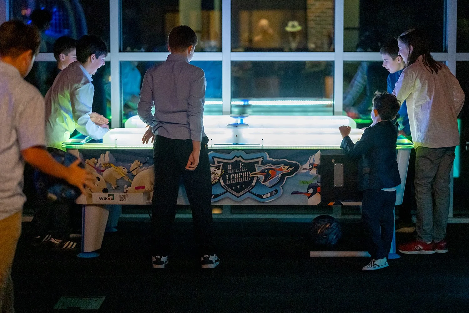 Children and adults playing air hockey indoors at night, with a large window in the background showing reflections and interior lights.