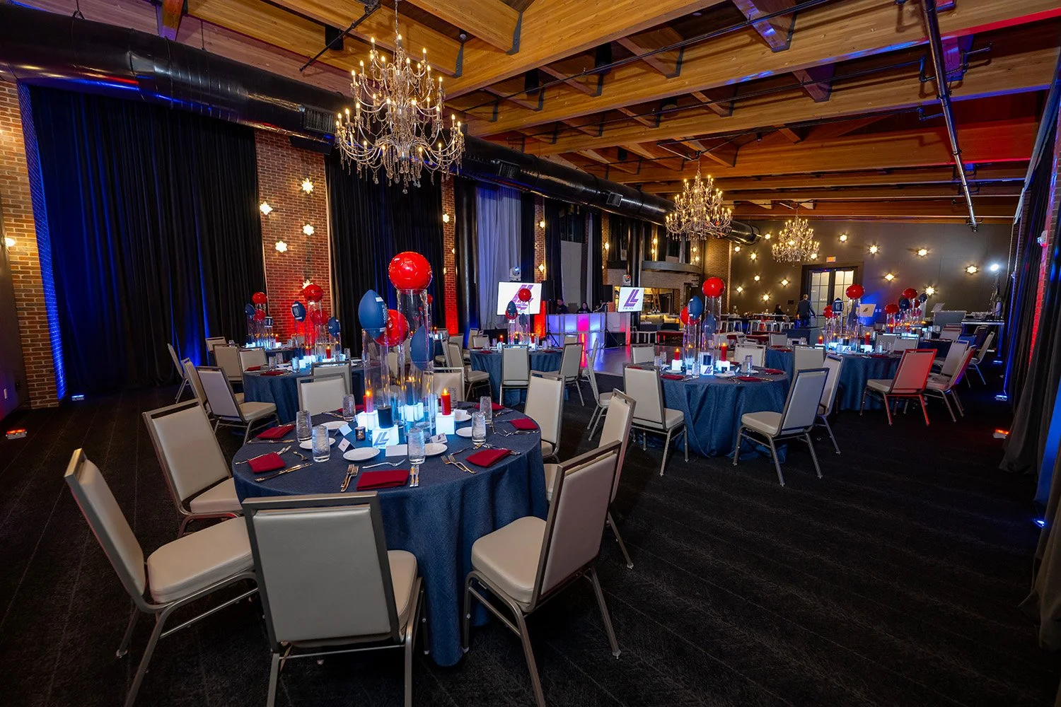 Elegant event space with round tables decorated with blue tablecloths, red and blue balloon centerpieces, and candles. Chandeliers hanging from a wooden ceiling, with brick and dark walls adorned with small lights, creating a festive atmosphere. A DJ