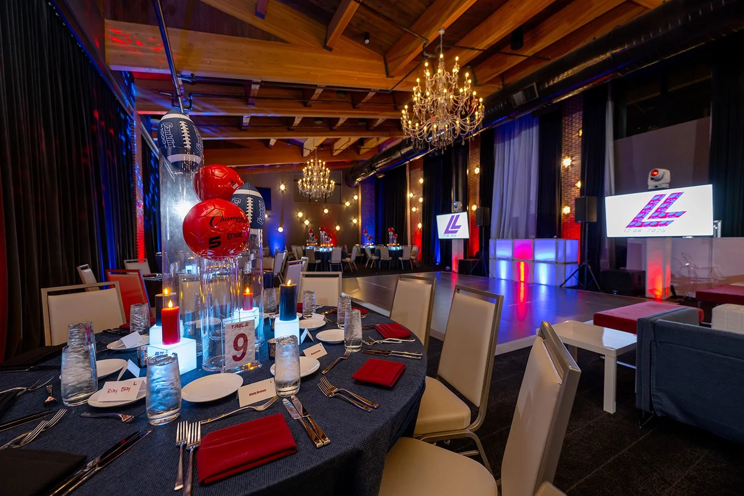A decorated event space with a round table set for a meal, featuring red and black napkins, water glasses, plates, and candles. In the background, there is a stage with large screens, chandeliers, and lighting that suggests a celebration or party, po