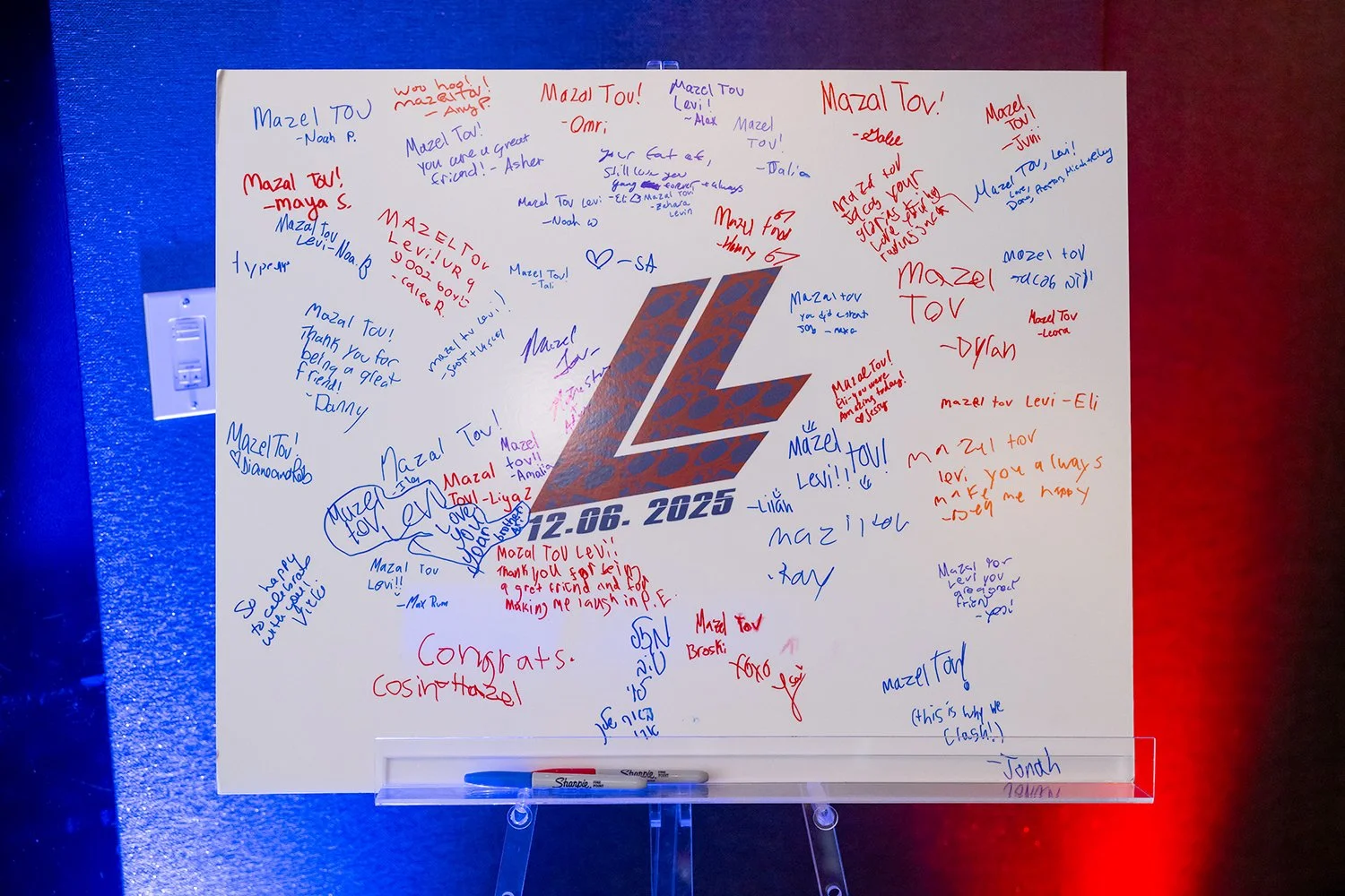 White poster with a large stylized 'L' logo in the center, surrounded by handwritten messages in red and blue ink. The messages include congratulations and well wishes, with some mentioning 'Mazal Tov' and the date 12.06.2025. The poster is mounted o