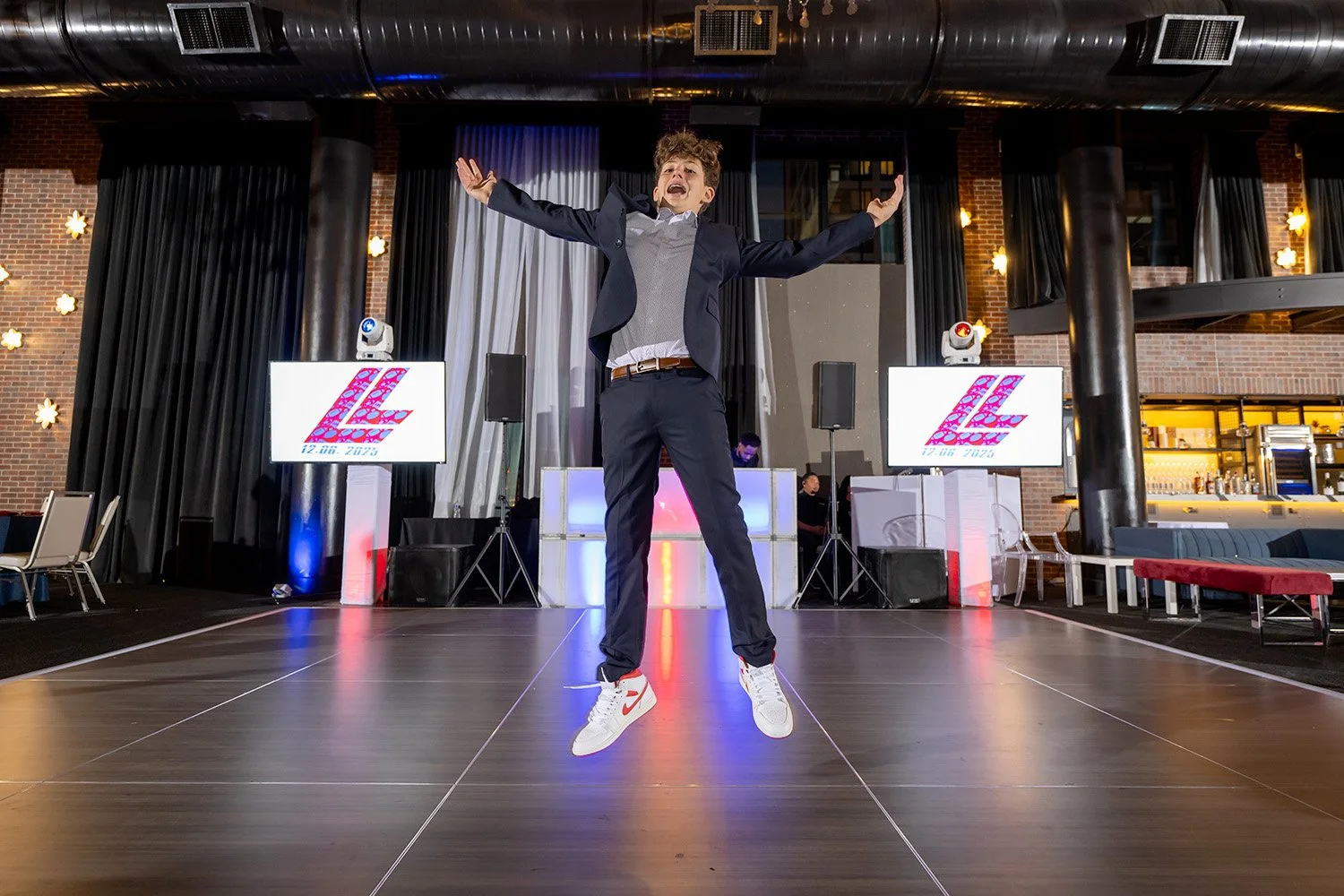 A young man in a suit and Nikes jumps in the air with arms spread wide on a dance floor at an event venue, with DJ equipment and two screens displaying the text 'LZ' and a date behind him.