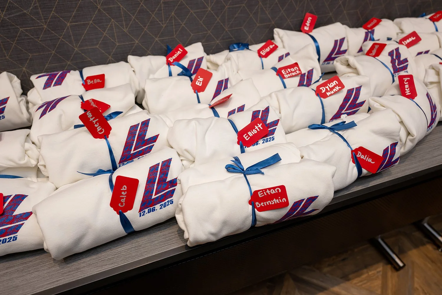 Multiple white fabric-wrapped items tied with blue ribbon, each labeled with a red tag displaying a name, arranged on a table, with a logo and date printed on the fabric.