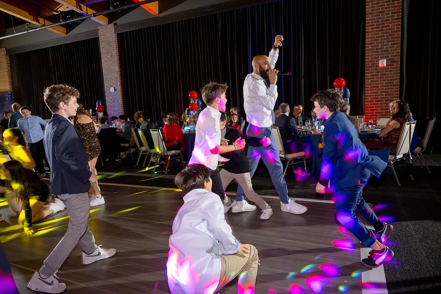 A DJ or singer performs on a stage at a party or event with guests dancing in front, some sitting at tables, colorful lights, and balloons in a decorated venue with brick walls.