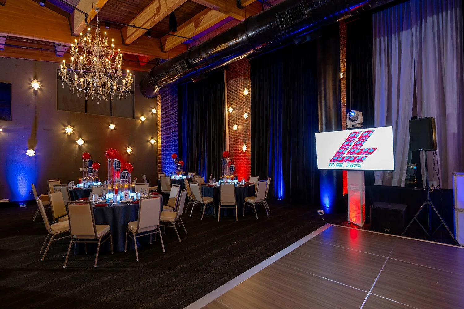 Event venue with decorated tables, a chandelier, blue and red lighting, a large screen displaying a logo with the initials 'L' and 'E', and a dance floor area.