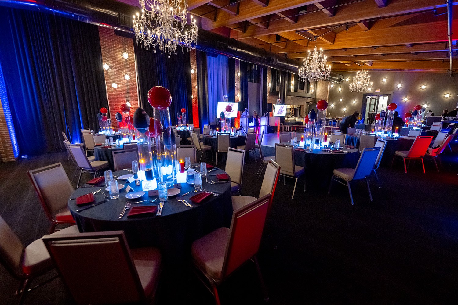 Elegant banquet hall decorated for a formal event with round tables covered in black tablecloths, red napkins, and tall centerpieces with red and black balloons. The hall features chandeliers, blue accent lighting, and a stage with screens in the bac