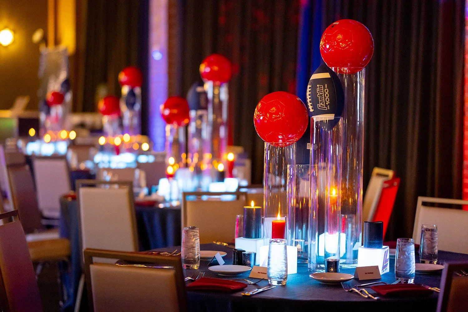 Elegant banquet table setting with tall glass vases containing red balloons, surrounded by lit candles, glasses, plates, and napkins, in a dimly lit event space with dark curtains.