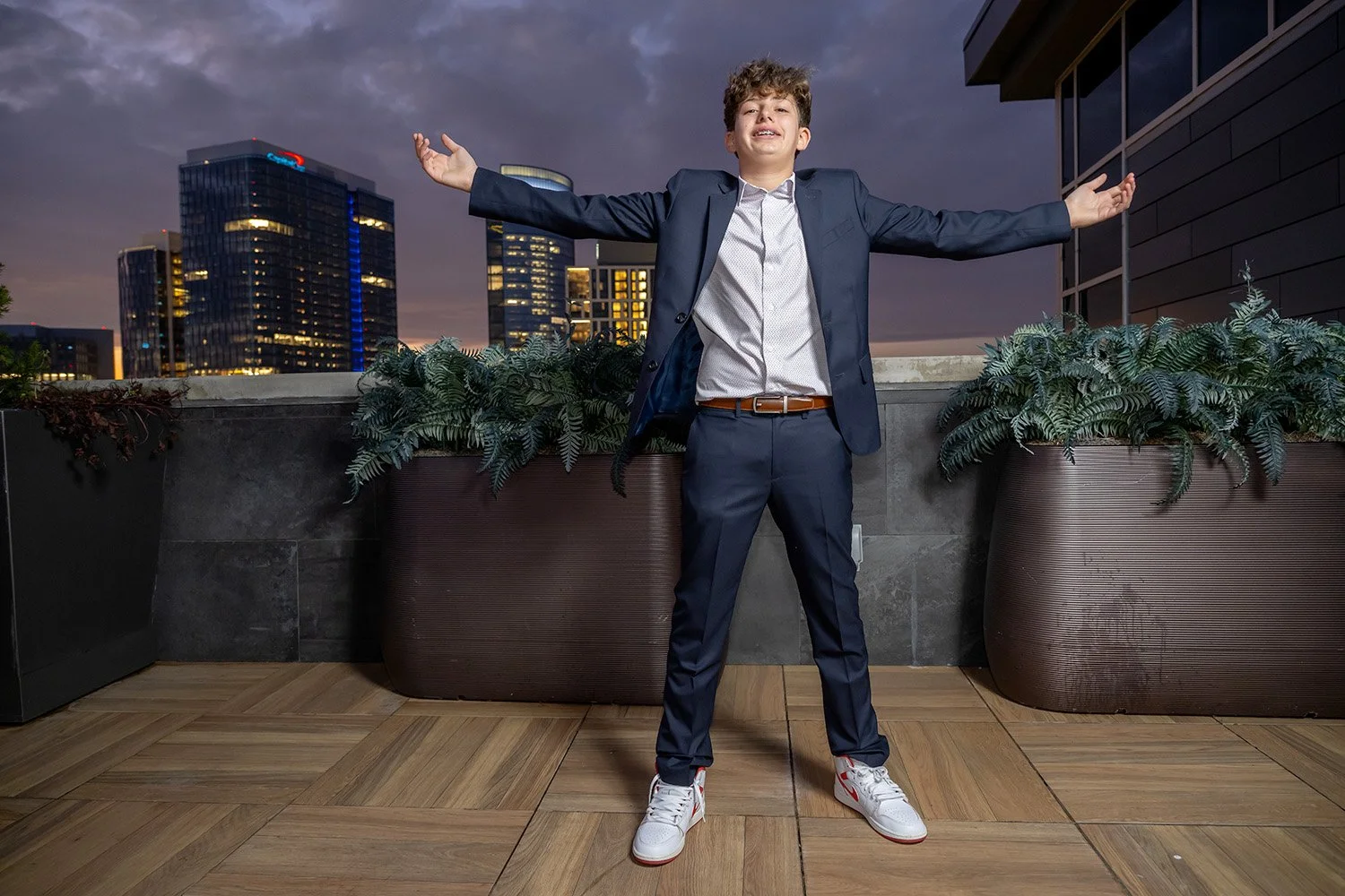 A young man in a suit and sneakers standing on a rooftop balcony at dusk with arms outstretched and a city skyline in the background.