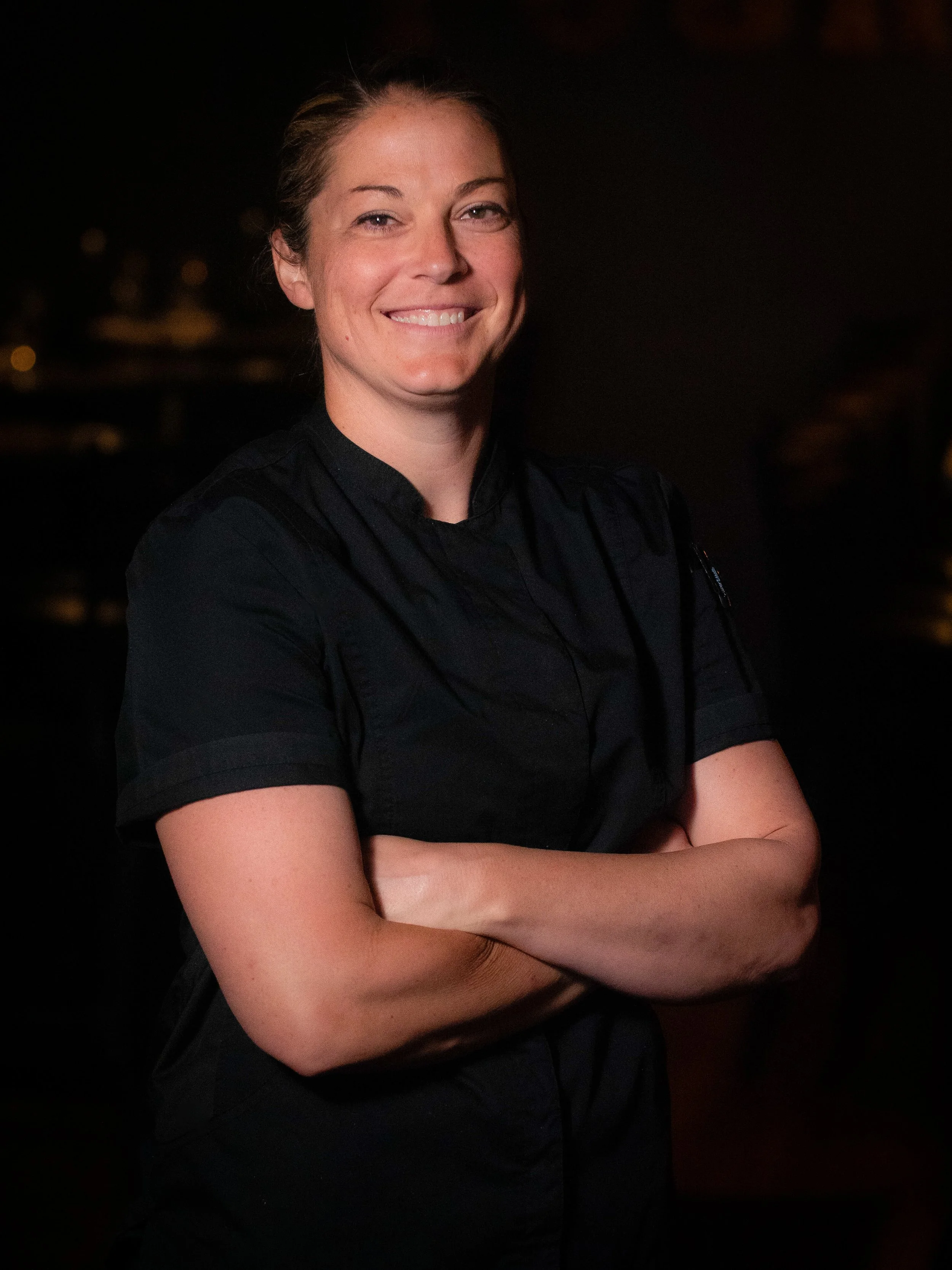 A woman in a black chef jacket smiling with arms crossed against a dark background.