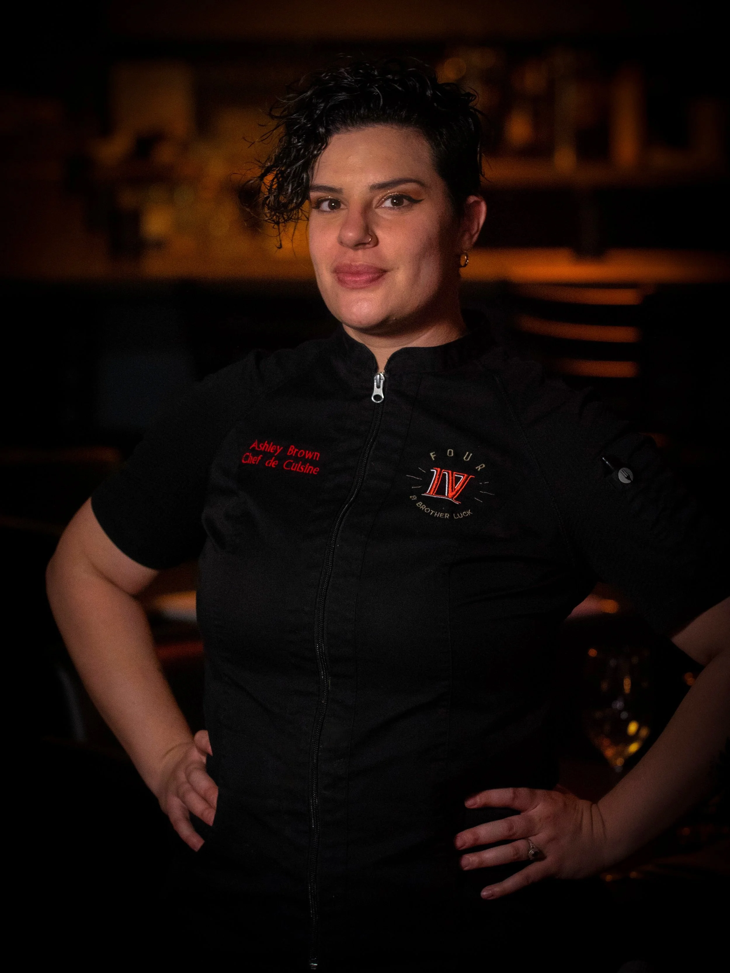 A woman with short, curly dark hair, tattoos on her arms, wearing a black chef's jacket with red embroidery that reads 'Ashley Brown, Chef de Cuisine' and a logo. She is standing with her hands on her hips in a dimly lit environment, possibly a restaurant or bar.