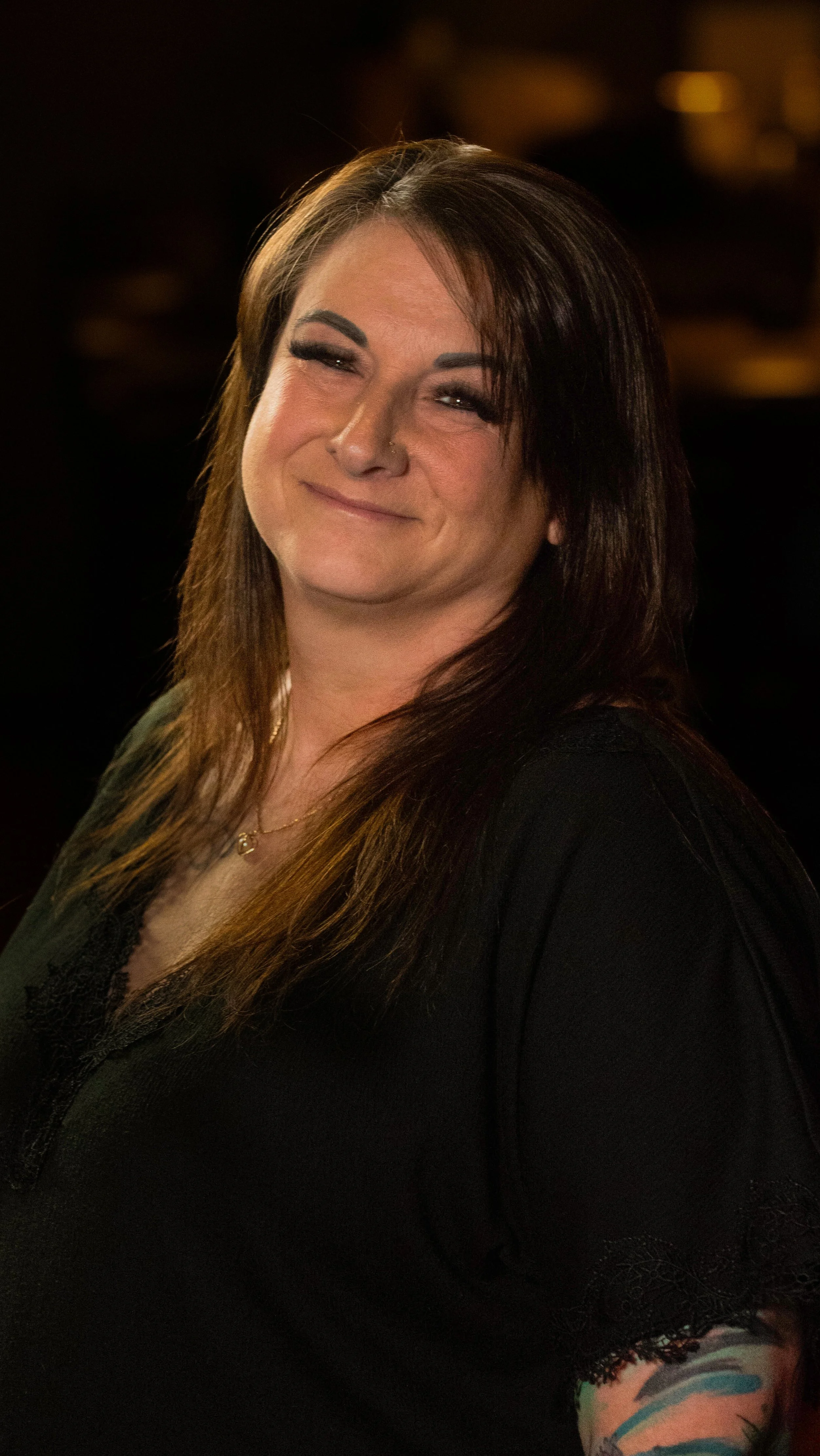 Portrait of a woman with dark brown hair, wearing makeup, a nose piercing, a black top with lace details, and a colorful tattoo on her arm, smiling with a dark blurred background.