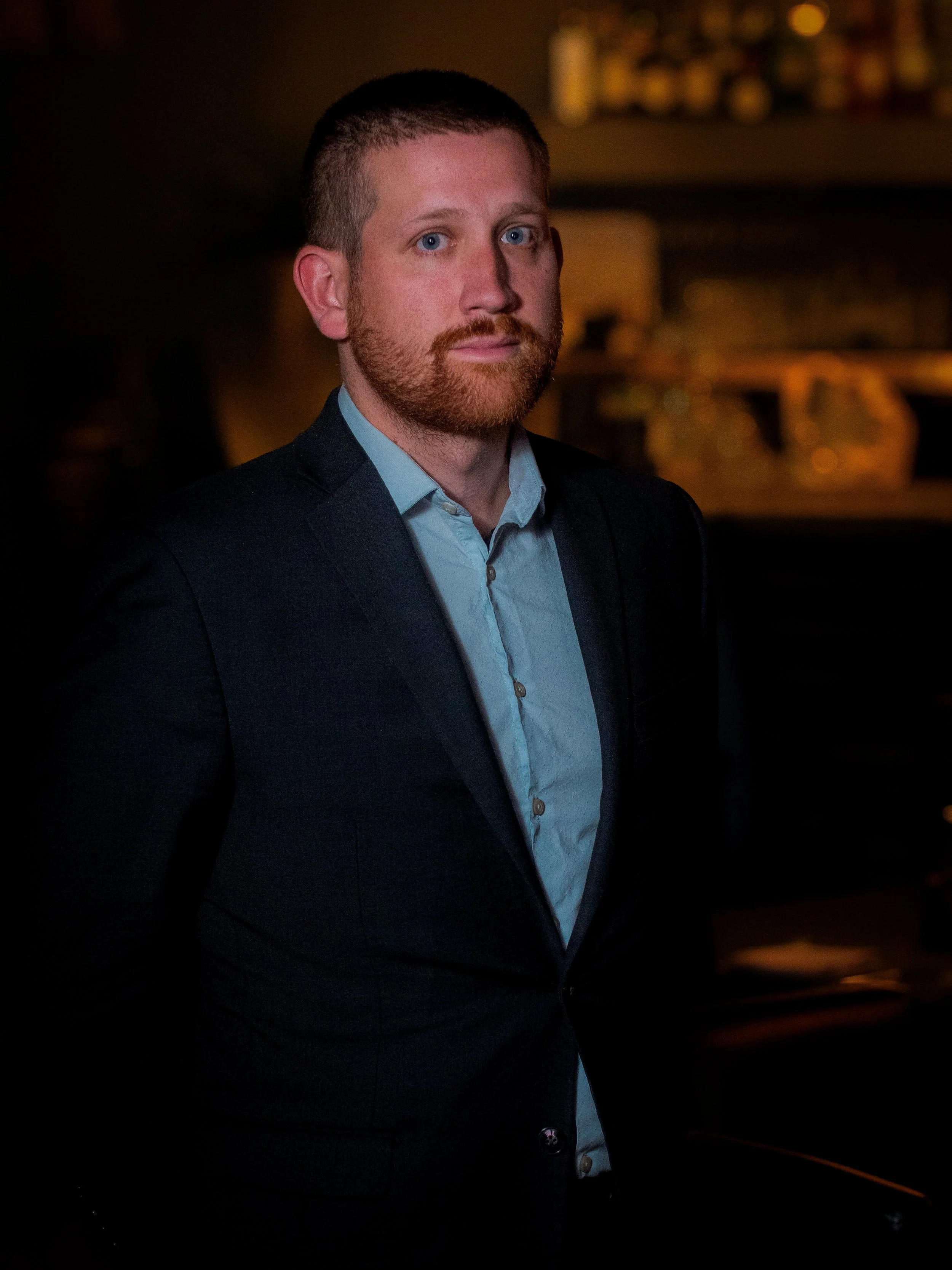 A man with blue eyes, red beard, and short hair, dressed in a dark suit and light blue shirt, standing in a dimly lit room.