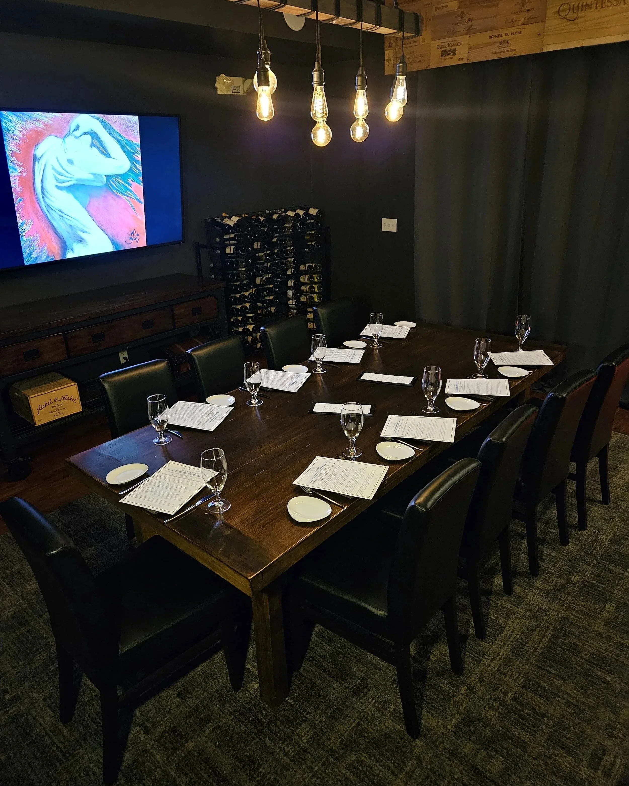 A dining room with a long wooden table set for six, with wine glasses, cloth napkins, and small potted plants, in a restaurant or cafe setting.
